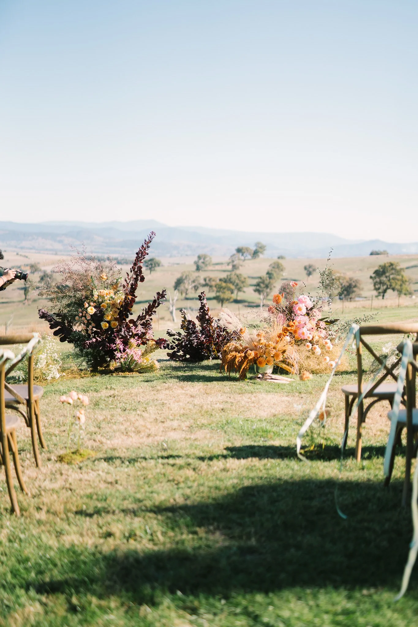 Outdoor wedding setup with floral arrangements, chairs, and a scenic landscape with rolling hills and trees under a clear blue sky.
