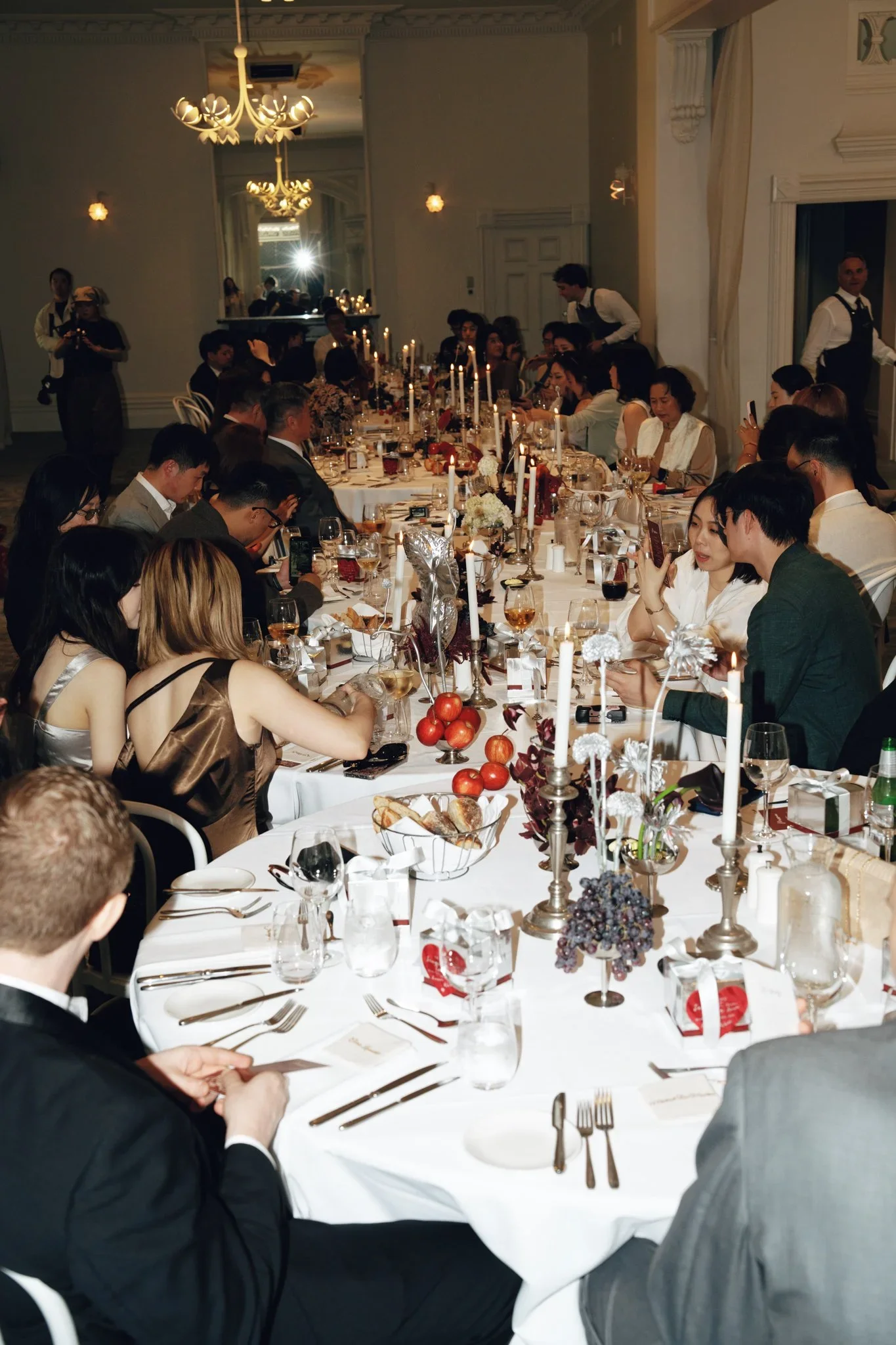 A formal banquet with guests seated around a long, decorated table. The table is set with white linens, candles, and floral centerpieces. People are engaged in conversations and using their phones. The room has elegant chandeliers and ornate decorati