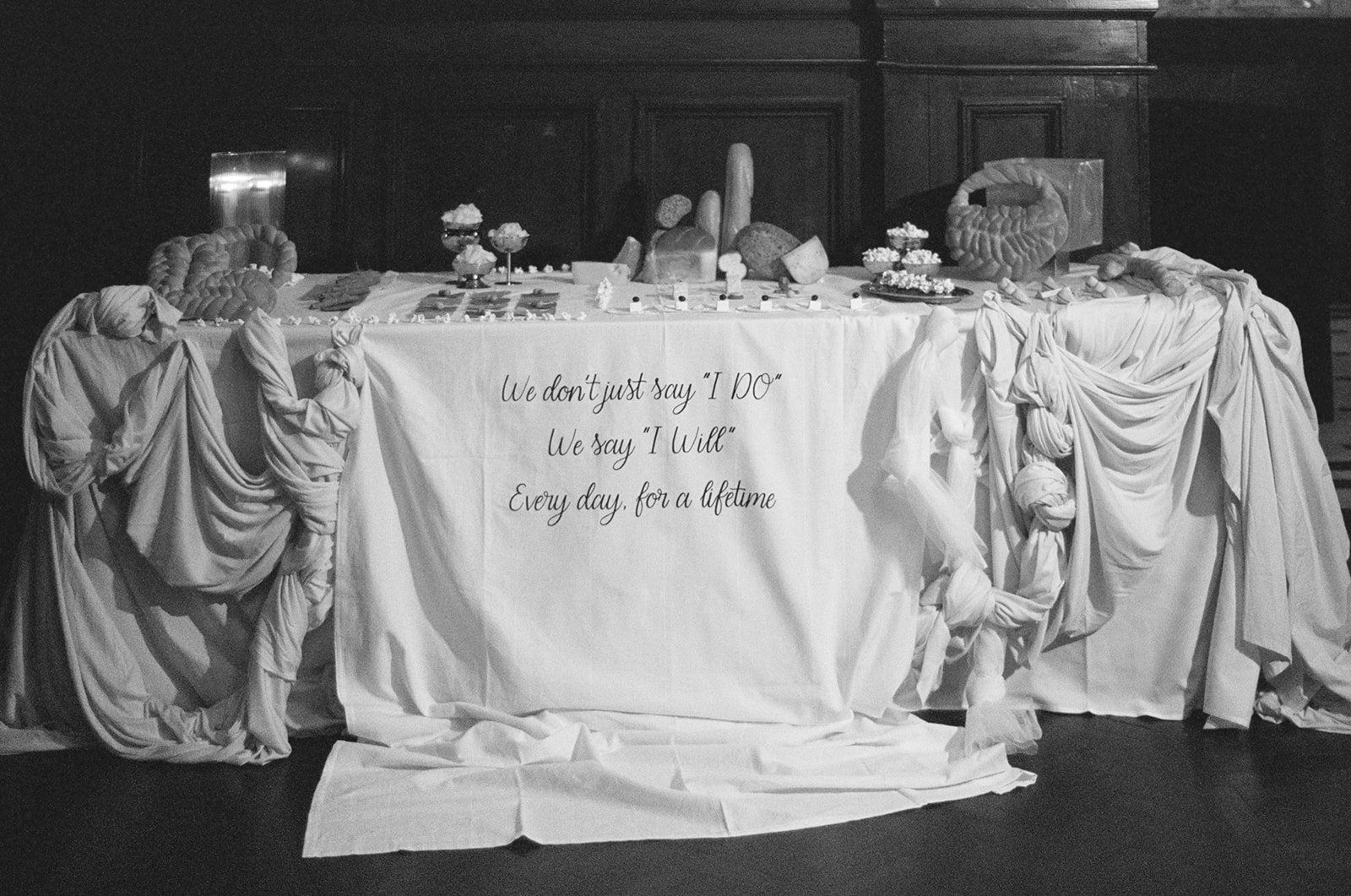 A table decorated with white drapes and carvings, displaying various objects including baked goods, bread, and small figurines. A quote on the front reads: 'We don't just say 'I DO' We say 'I Will' Every day, for a lifetime.'