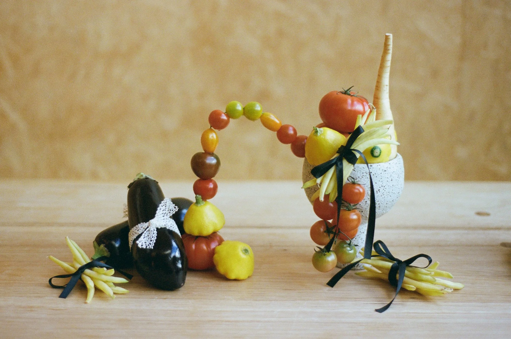 A colorful arrangement of fruit and vegetable sculptures, including an eggplant with a lace bow, a cherry tomato, a lemon, and a parsnip, all arranged in a white speckled pot with black ribbons, on a wooden surface against a beige background.