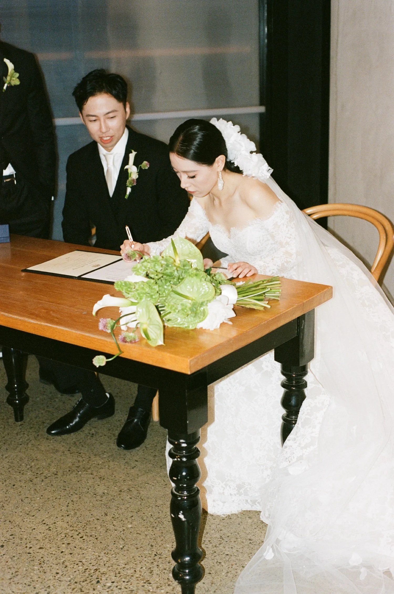 A bride with dark hair in an updo and wearing large earrings signs a document at a wedding reception. She is dressed in a white lace wedding gown and is leaning over a wooden table with a green floral arrangement. A groom in a black suit with a bouto