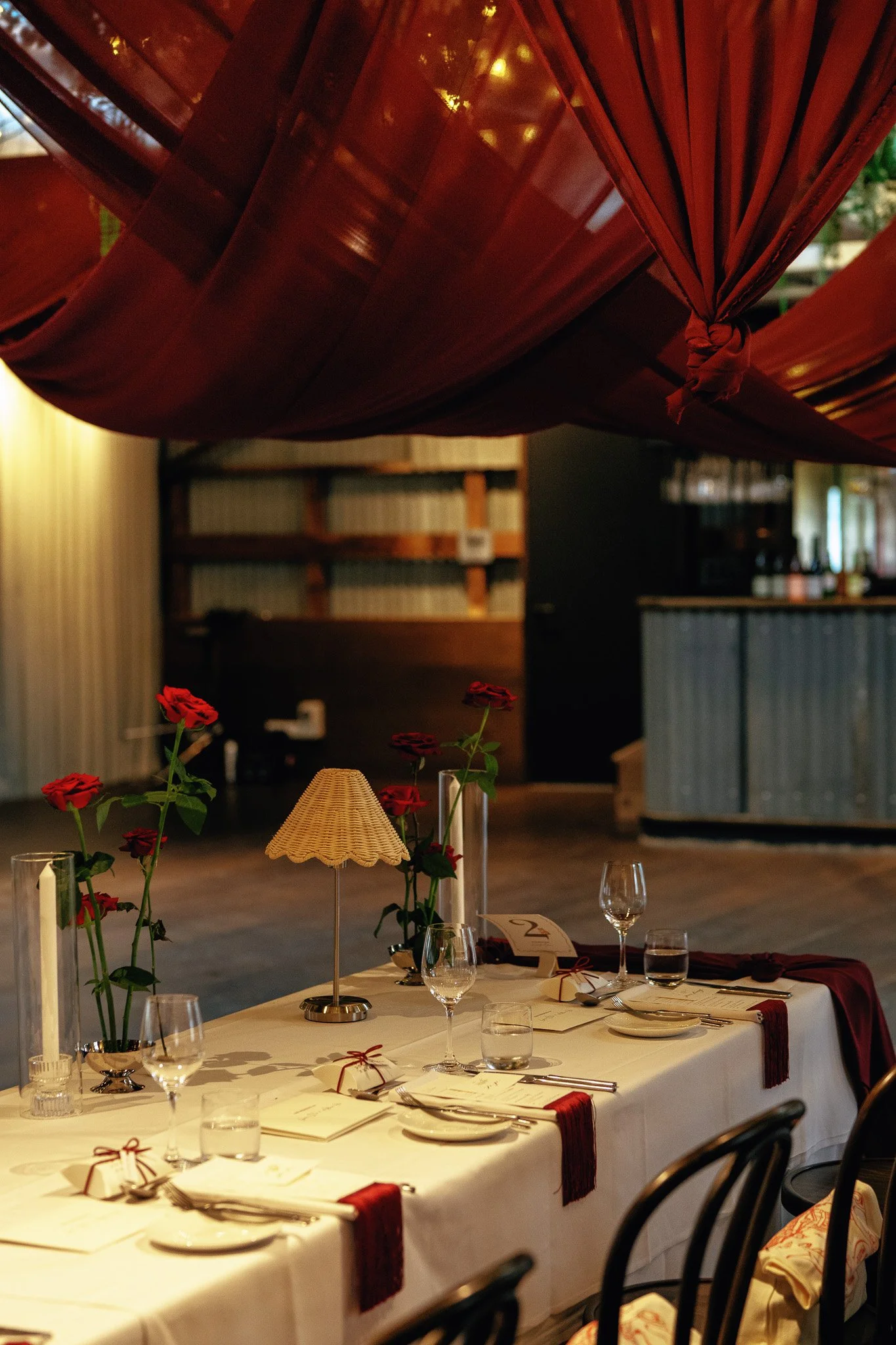 A dining table set with white tablecloth, glasses, silverware, and small wrapped gifts, decorated with red roses and a small lamp, in a warmly lit restaurant or event space with wooden and paneled walls, and a large red canopy overhead.