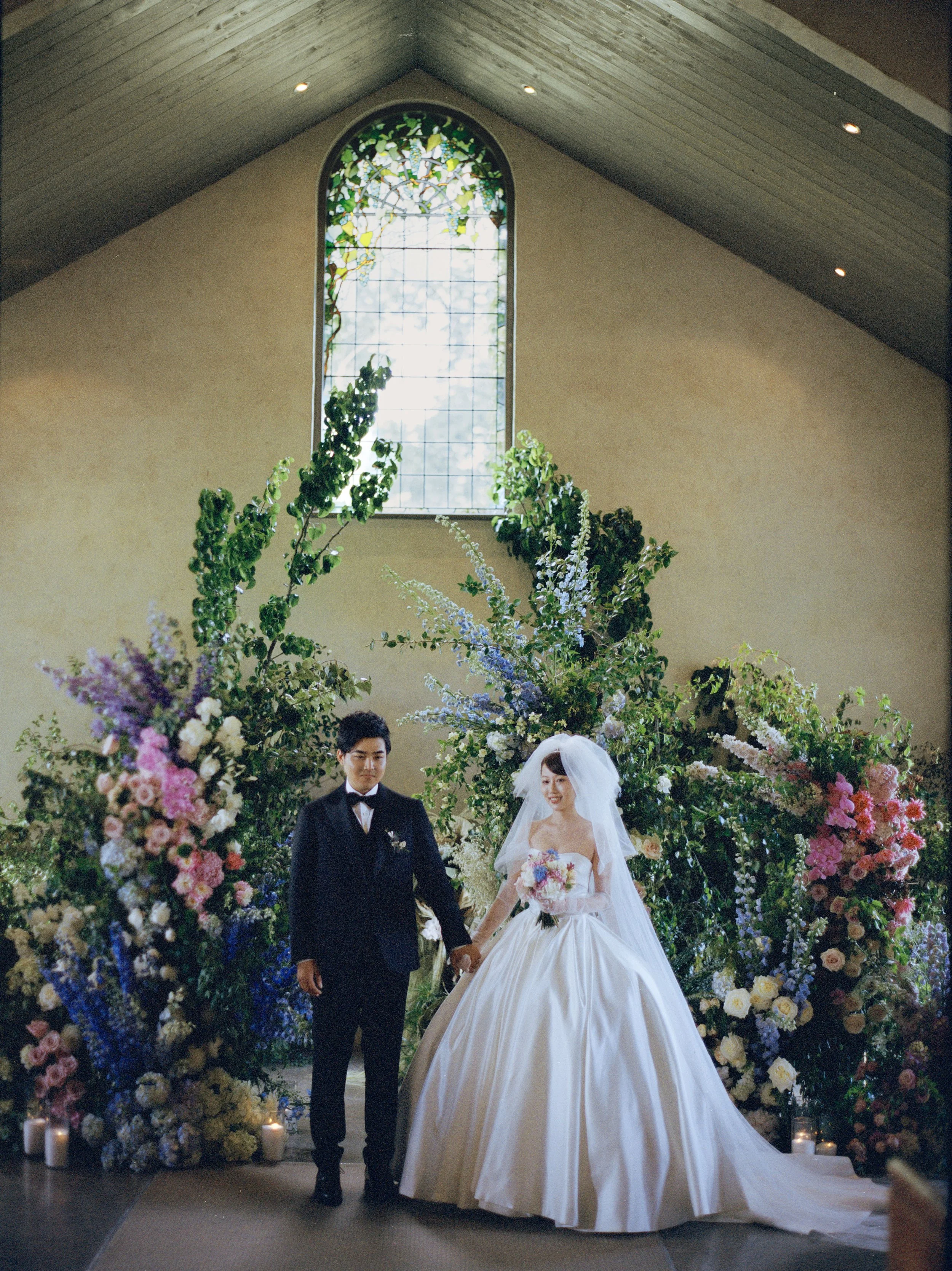 A newlywed couple holding hands during a wedding ceremony inside a church or chapel, surrounded by a large floral arch with various colorful flowers and greenery, a stained glass window behind them, and candles at the base of the floral arrangement.