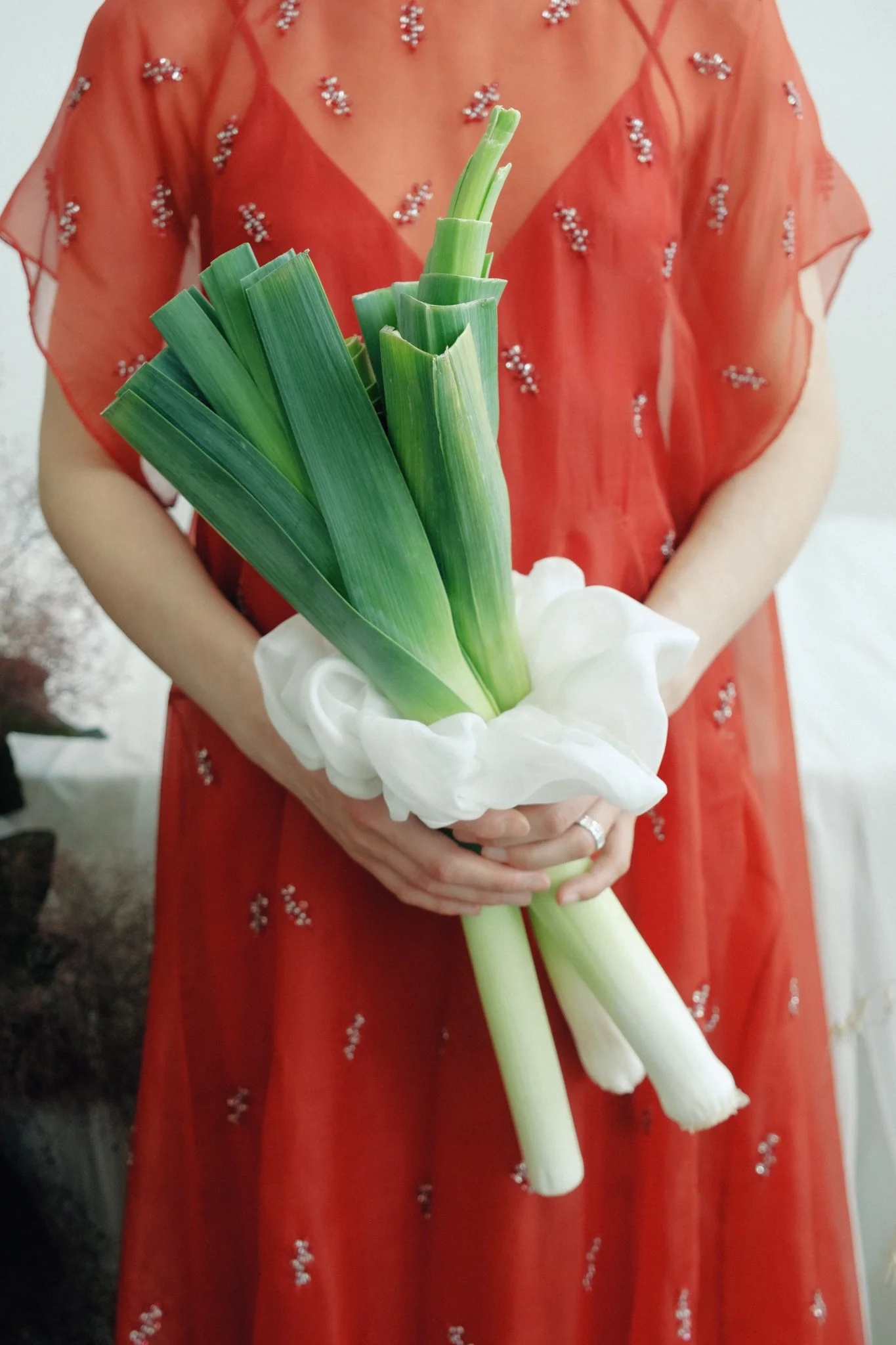 A person in a red dress decorated with silver sequins is holding a bouquet of green and white flowers, including leeks and calla lilies.