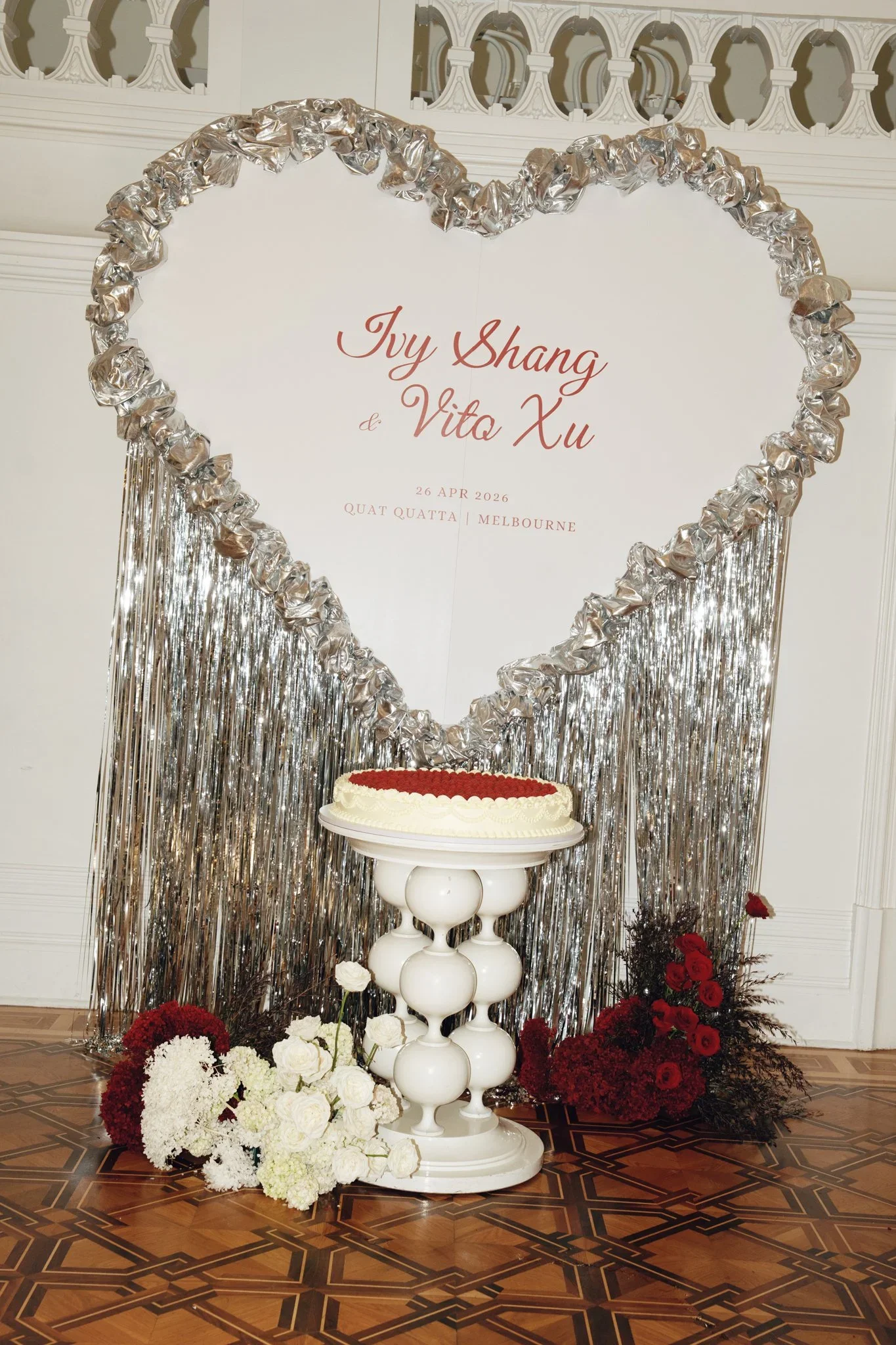 A wedding celebration setup with a heart-shaped metallic decoration frame, a white sign with red cursive text displaying the names 'Jiy Shang' and 'Vito Xu,' the date April 26, 2026, and the location 'Quat Quatta Melbourne.' There is a white tiered t