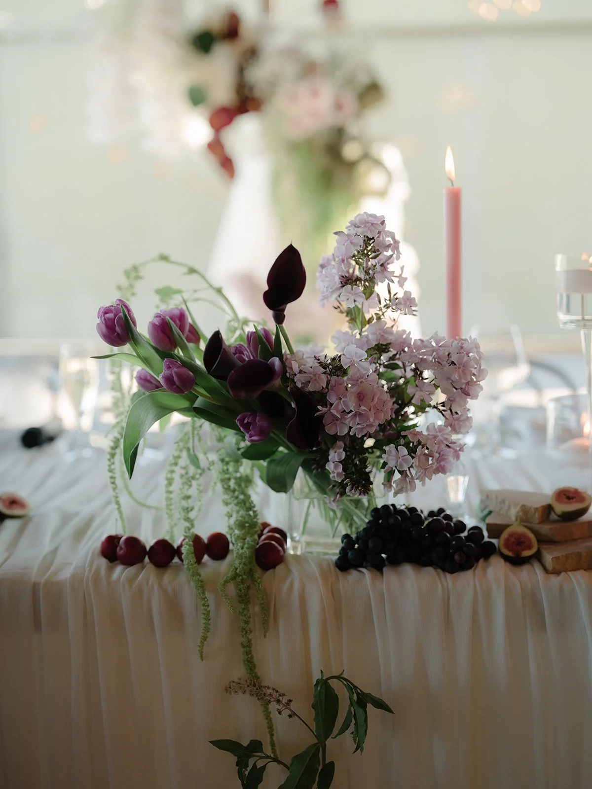 Elegant floral centerpiece with pink tulips, dark purple calla lilies, white hydrangeas, and hanging greenery on a white tablecloth, with a pink candle in the background.