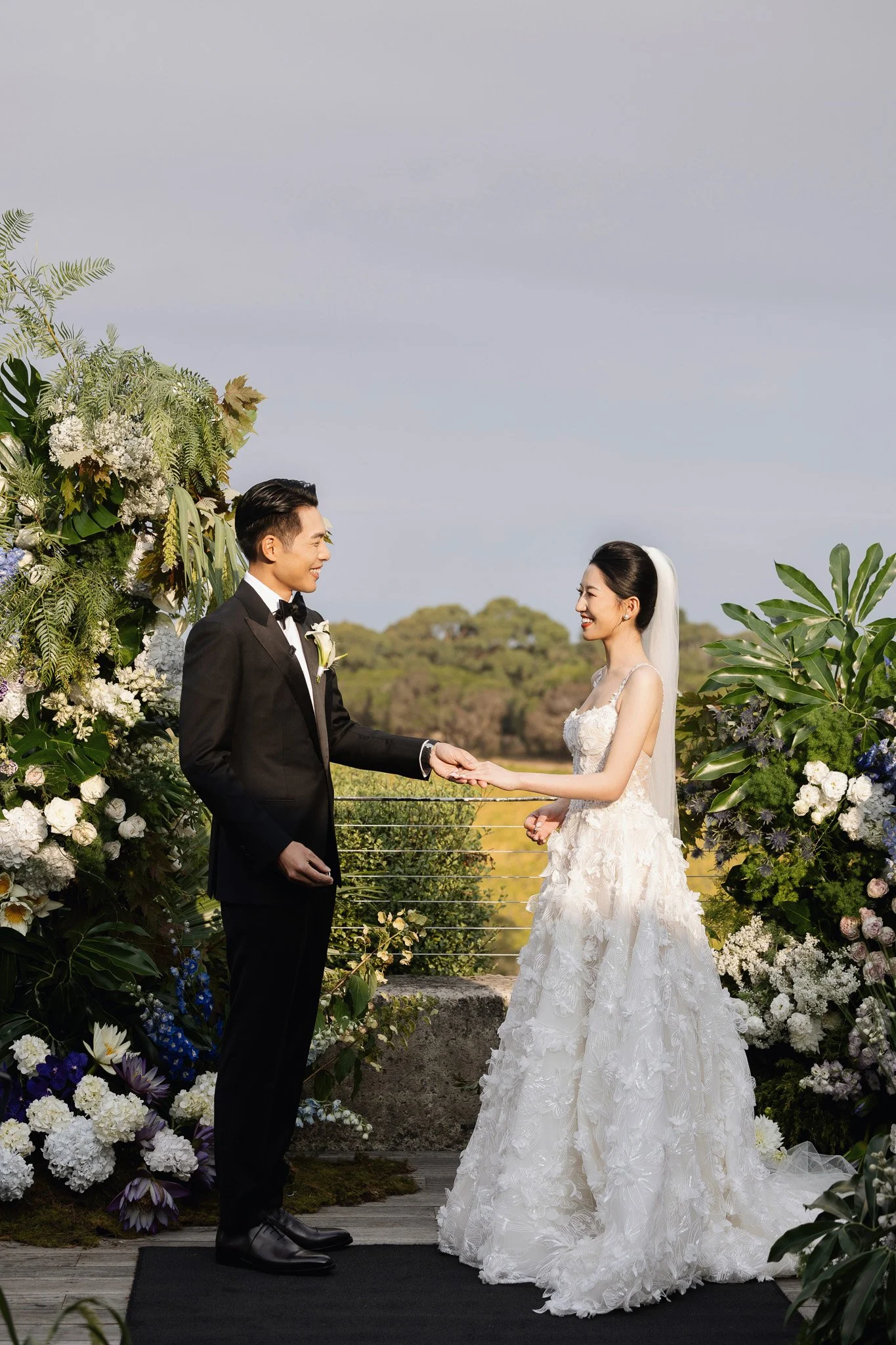 A bride and groom holding hands during their outdoor wedding ceremony, surrounded by lush green plants and white flowers, with a scenic landscape in the background.