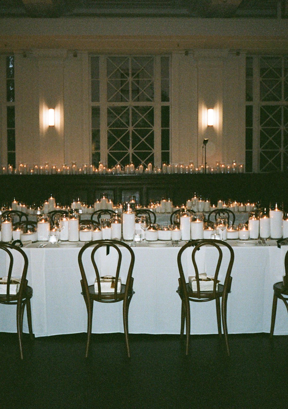 Elegant banquet table decorated with numerous white candles and string lights, set for an event in a dimly lit room with large windows and wall sconces.