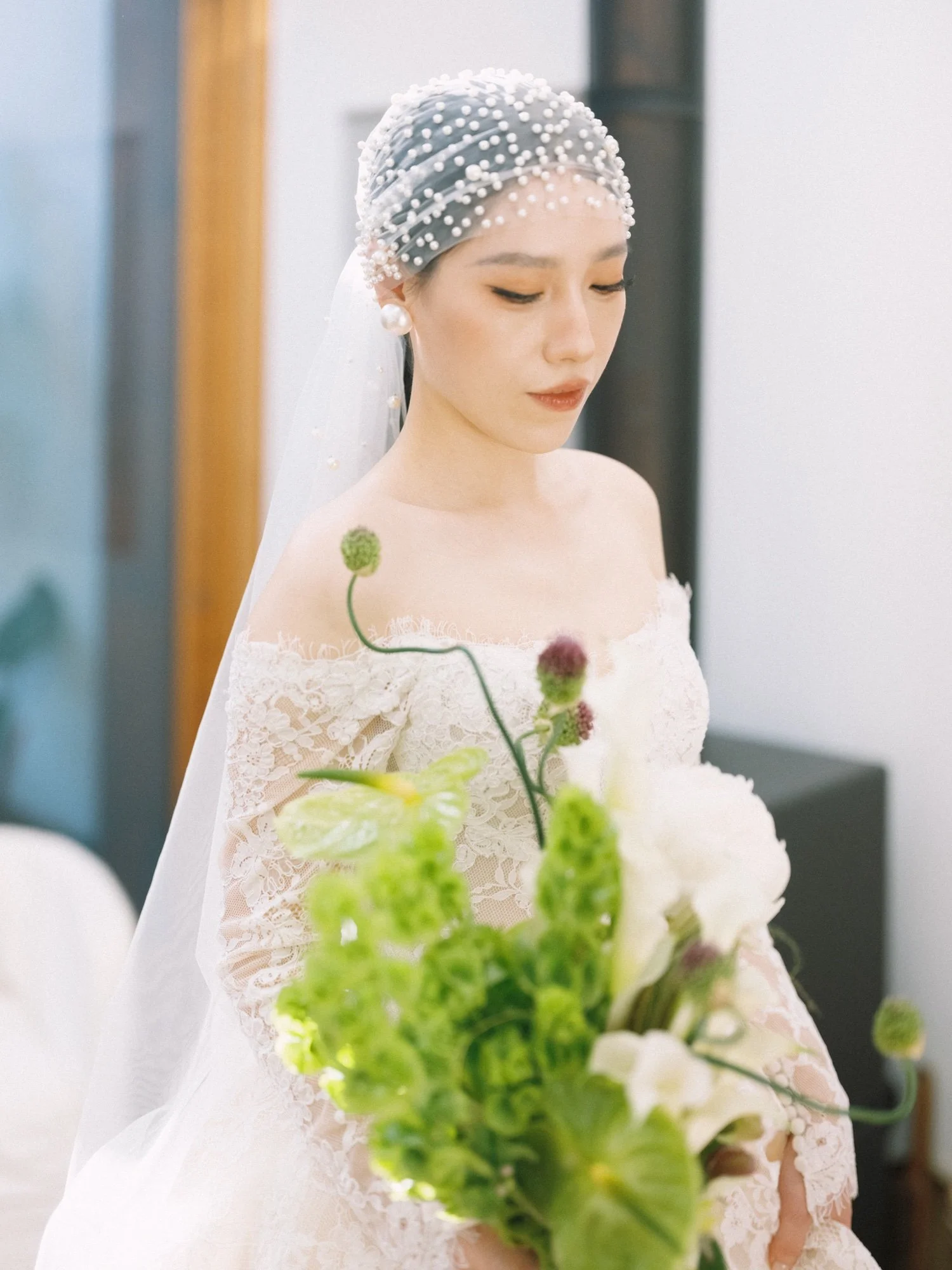 A woman in a white lace dress and pearl accessories is looking down, holding a bouquet with green and pink flowers, with a blurred background.