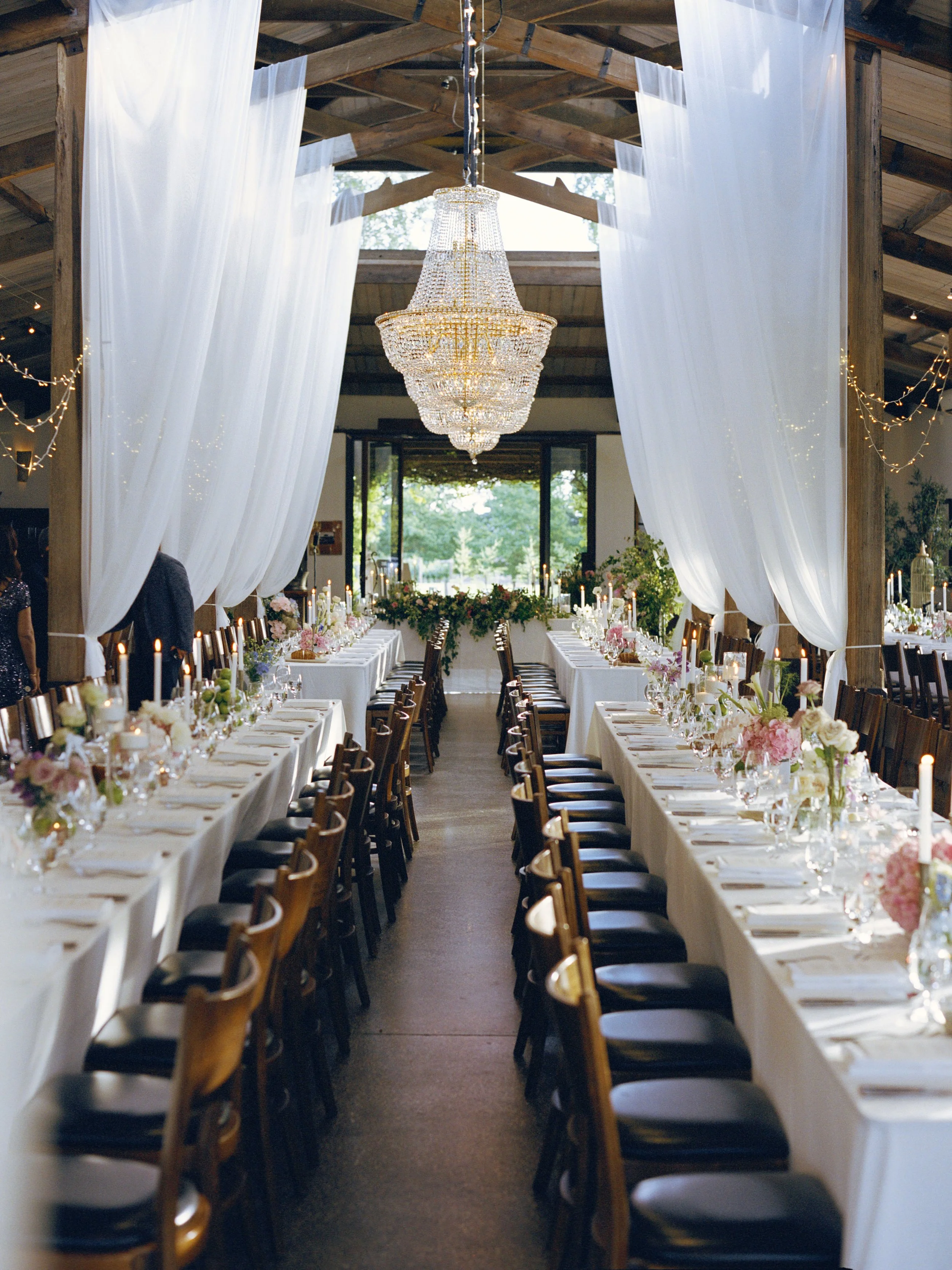 A long banquet table with white tablecloths arranged with flowers and candles, set up in a rustic barn with wooden beams, white drapes, and a large chandelier hanging from the ceiling, ready for a celebration or event.