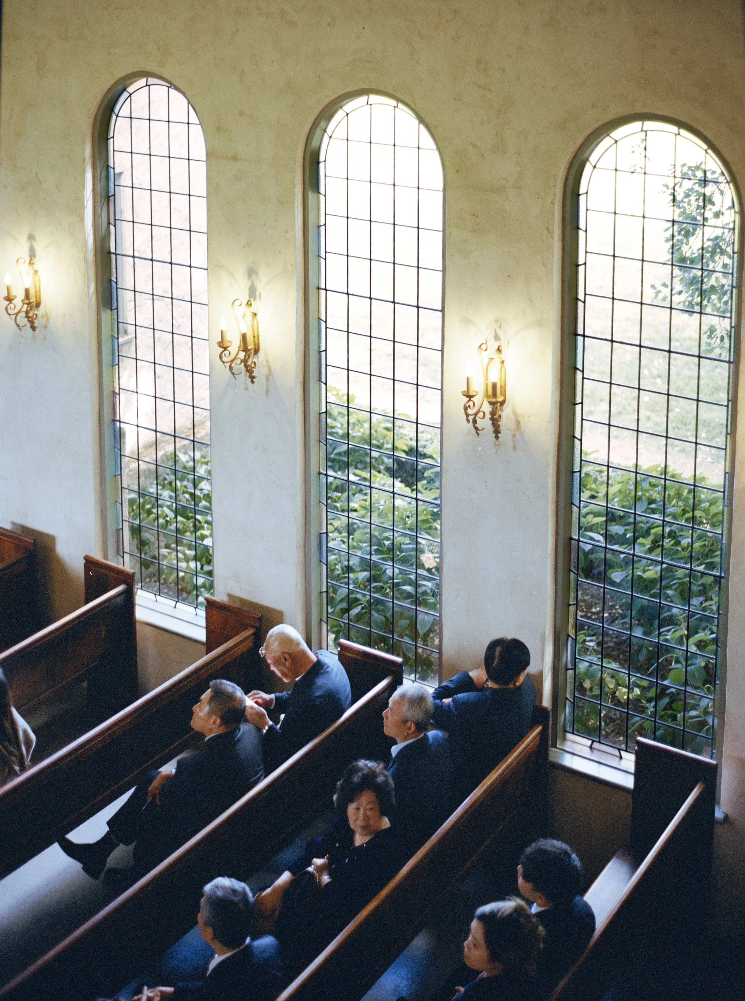 Inside a church with large arched stained glass windows, several people dressed in formal attire are seated in pews, attending a service, with greenery visible through the windows.