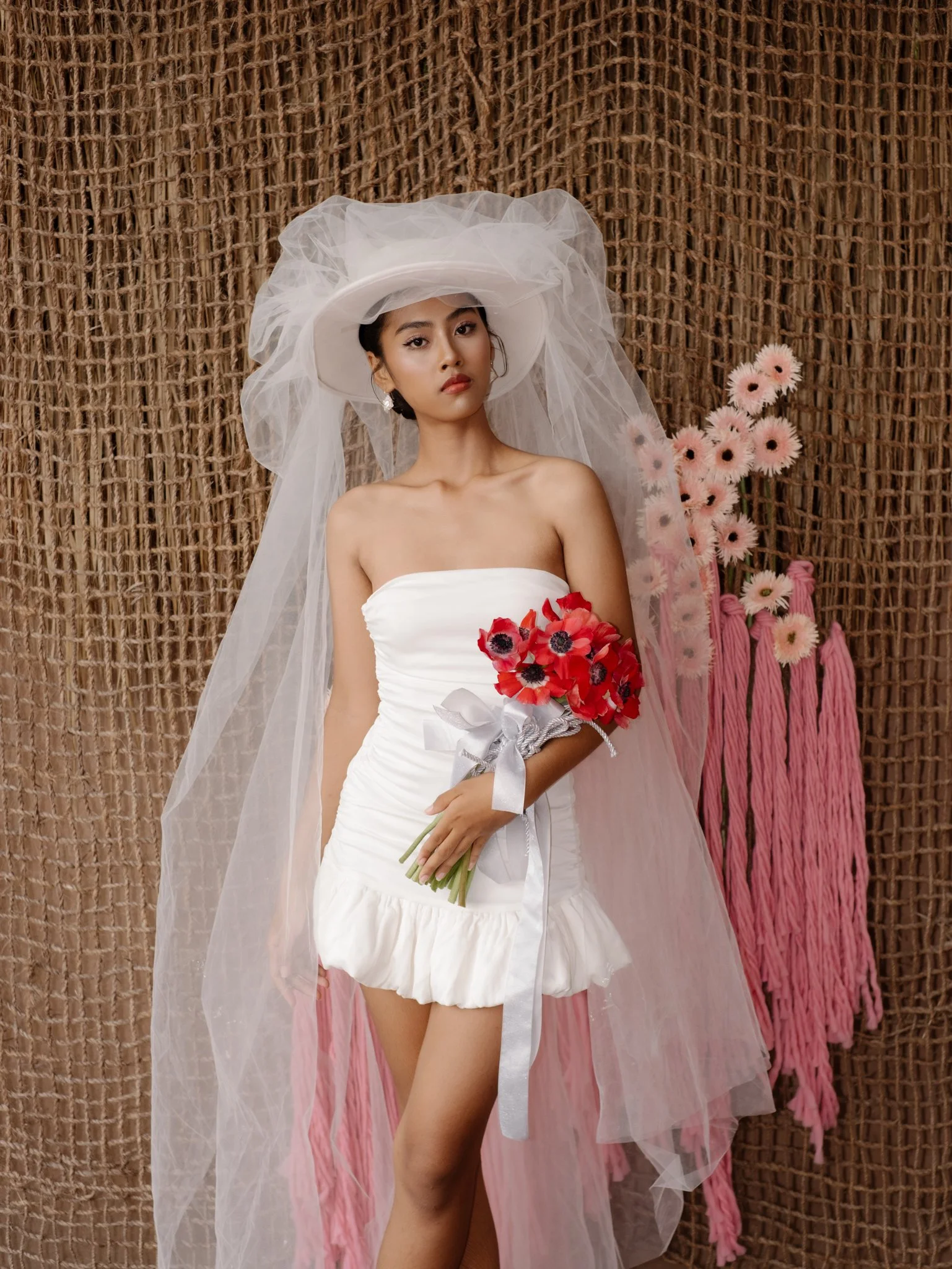 A woman in a strapless white dress holding a bouquet of red flowers with dark centers, standing in front of a textured woven background, wearing a large white hat adorned with sheer fabric tulle and earrings, with pink flowers and pink tassels on the