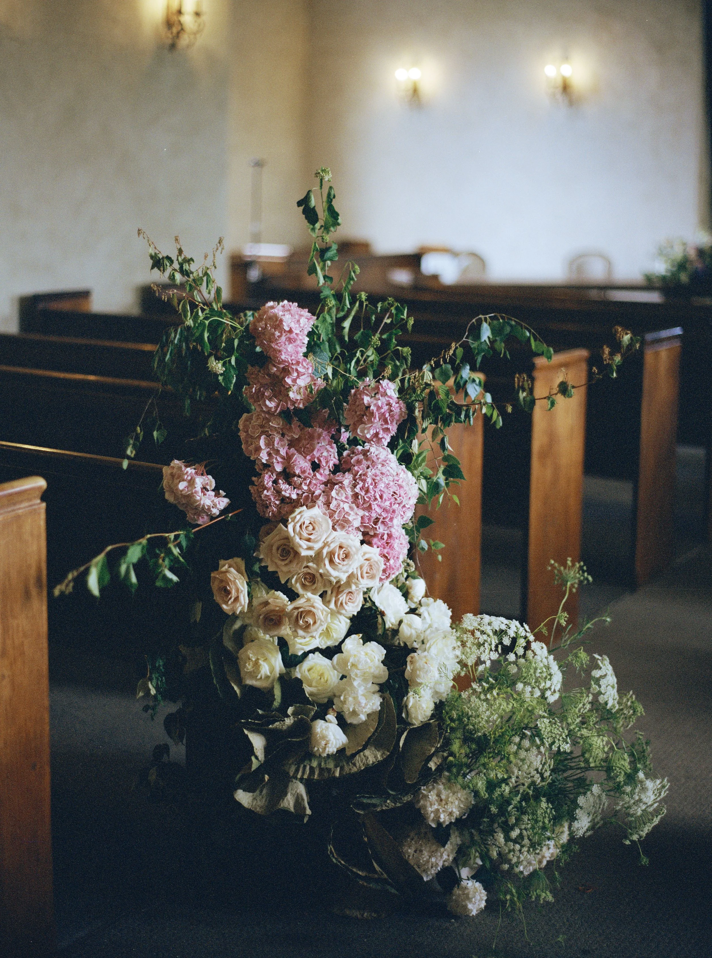 Arrangement of pink and white flowers, including roses and hydrangeas, placed on a dark surface in a room with wooden furniture and soft lighting.