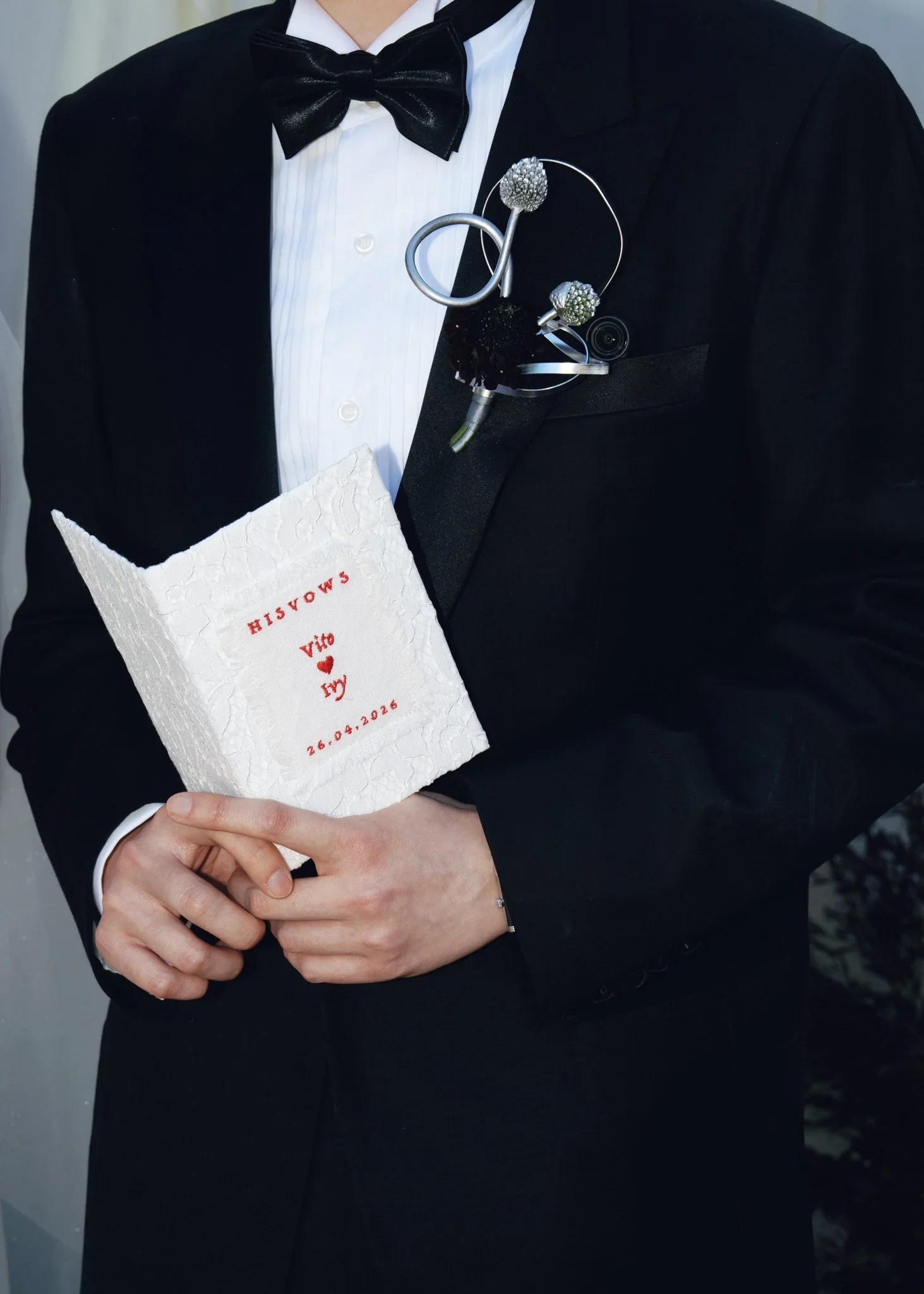 Person in a tuxedo holding a white textured card with red text and a heart symbol. The tuxedo features a black jacket, white shirt, and a black bow tie. A decorative silver lapel pin with intricate design is pinned to the jacket.