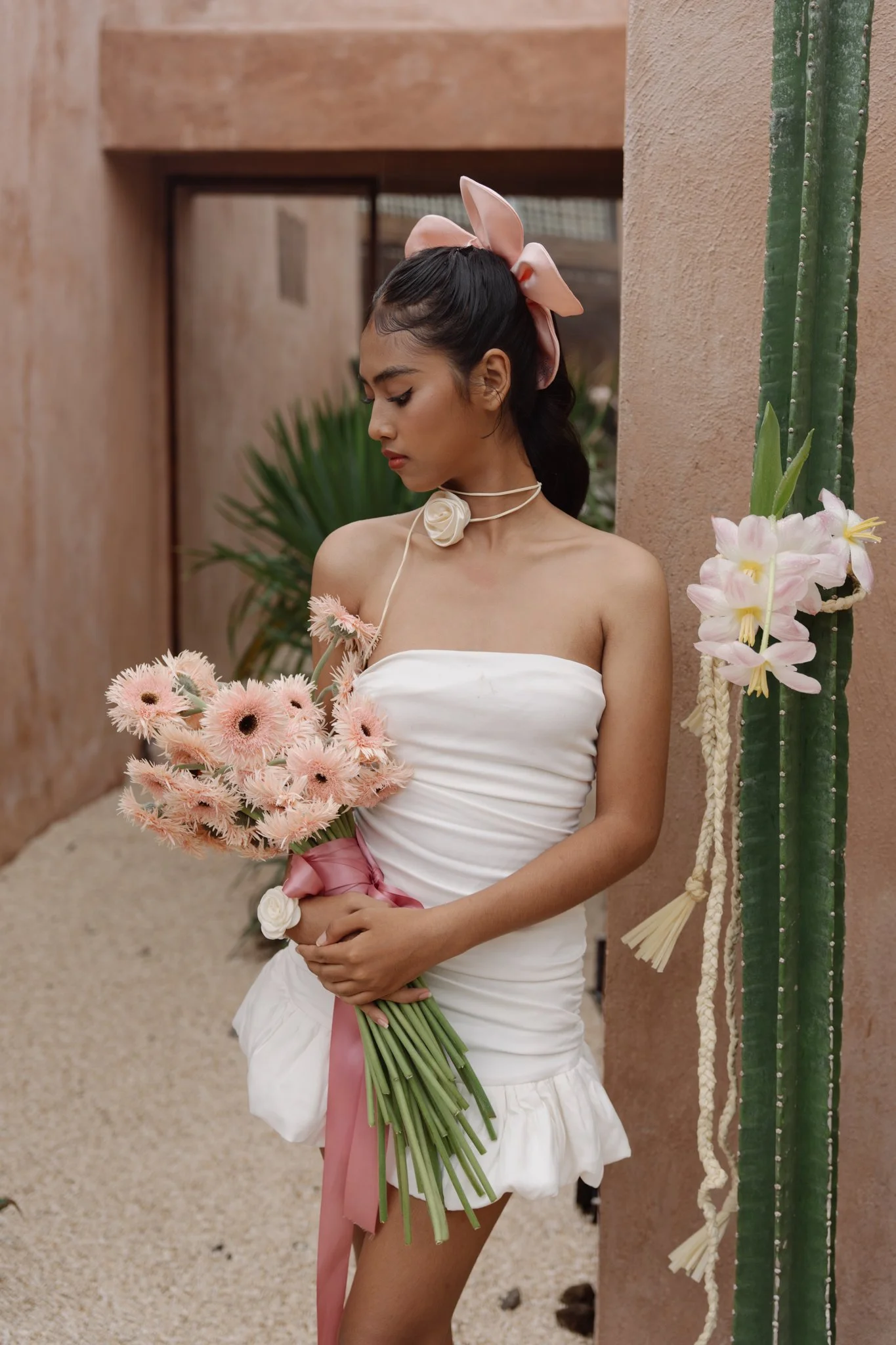 A young woman with dark hair styled in waves, wearing a pink bow headband and a white strapless dress with a ruffled skirt, holds a bouquet of pink flowers tied with a pink ribbon, standing next to a tall cactus with pink flowers and a beige wall bac