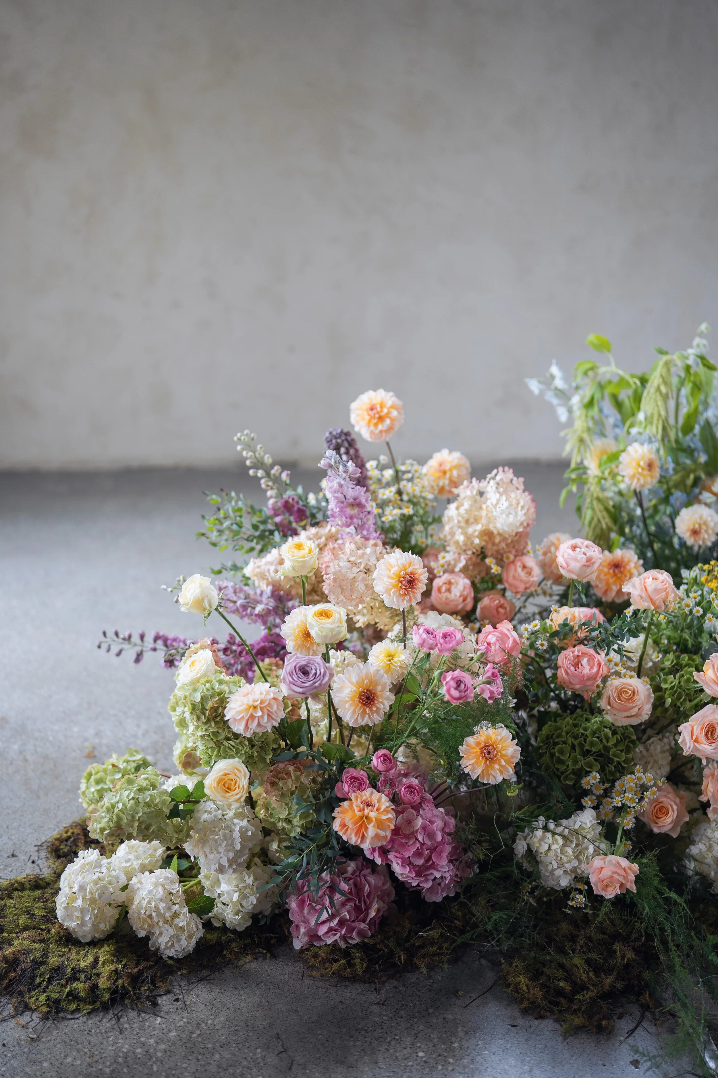 A colorful floral arrangement features roses, hydrangeas, dahlias, and other flowers in pastel shades of pink, peach, purple, and white, placed on a gray surface with moss.