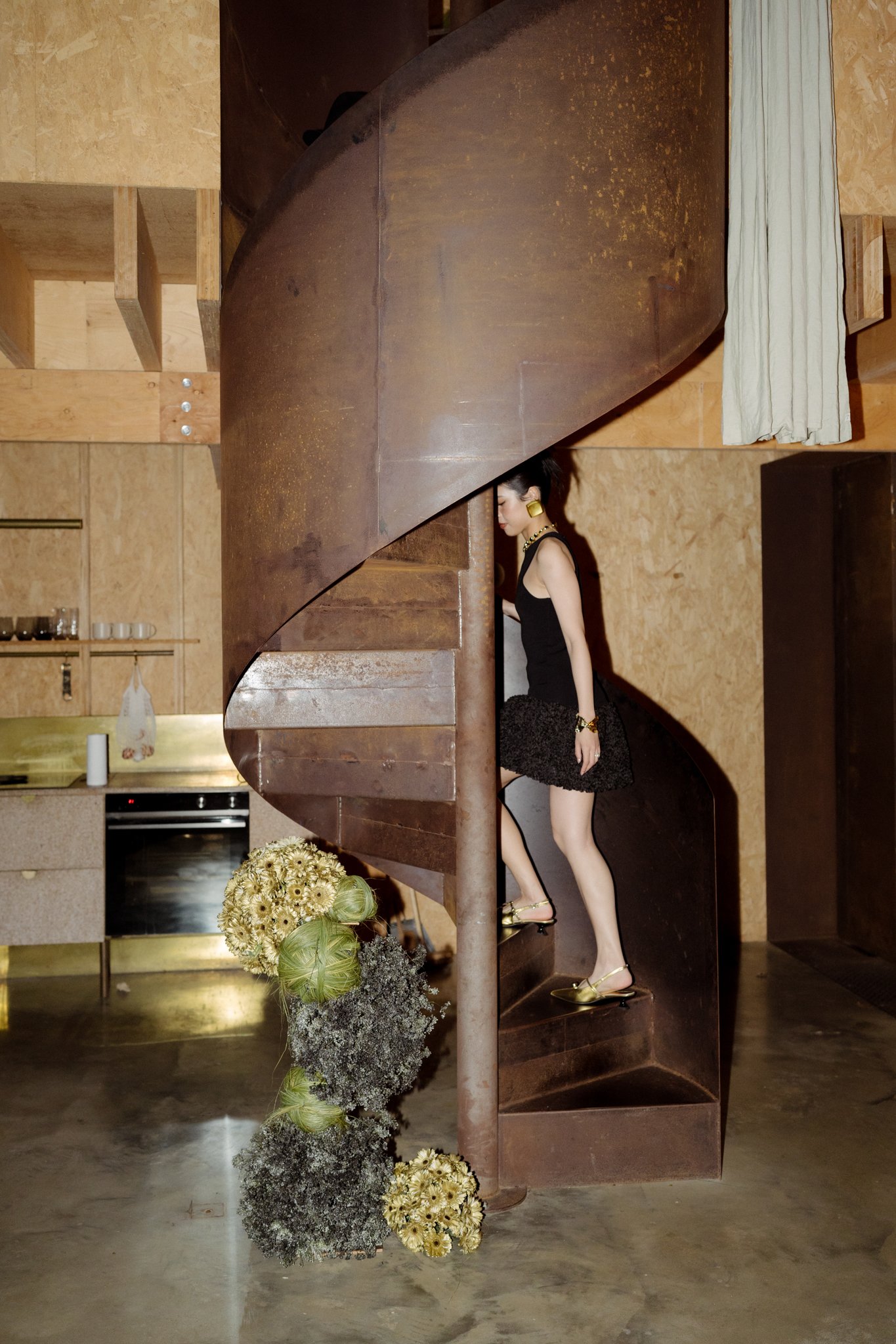 A woman in a black dress and gold high heels walking up a curved staircase made of wood and metal inside a room with wooden walls and kitchen appliances, and a floral arrangement below the staircase.