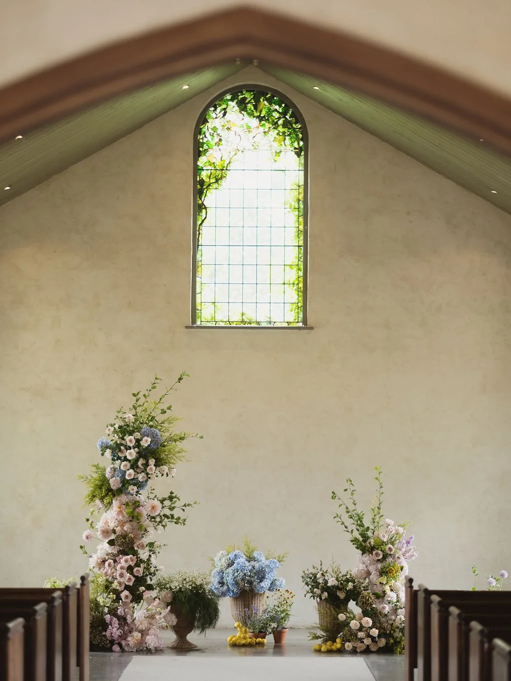 Inside a church with a high ceiling and a stained glass window at the front, decorated with multiple floral arrangements featuring pink, blue, and white flowers.