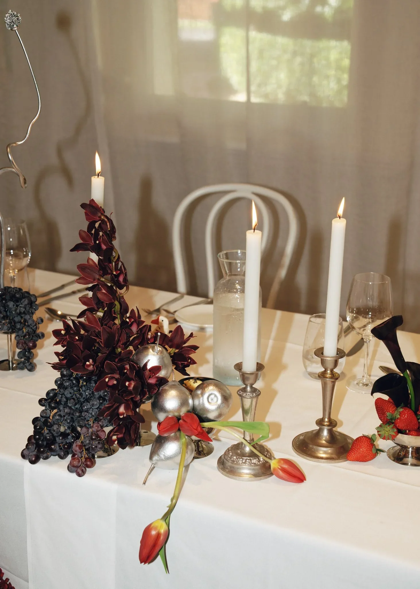 A table decorated with candles, flowers, berries, and ornaments, set for a special occasion or celebration, with chairs and a window in the background.