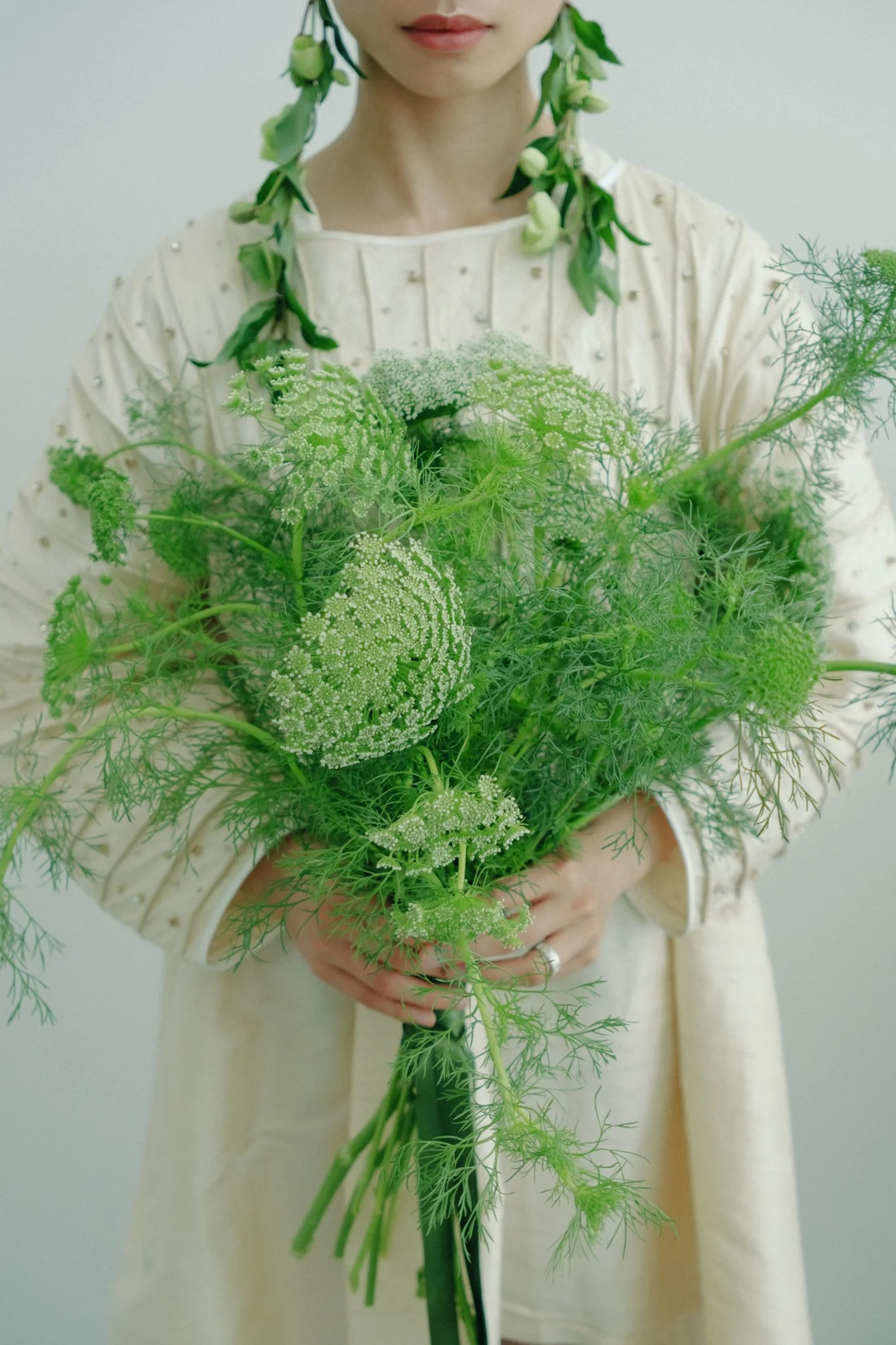 A woman dressed in a cream-colored outfit with pearl embellishments on her shoulders, holding a bouquet of green flowers and foliage. She is standing against a plain, light-colored background.