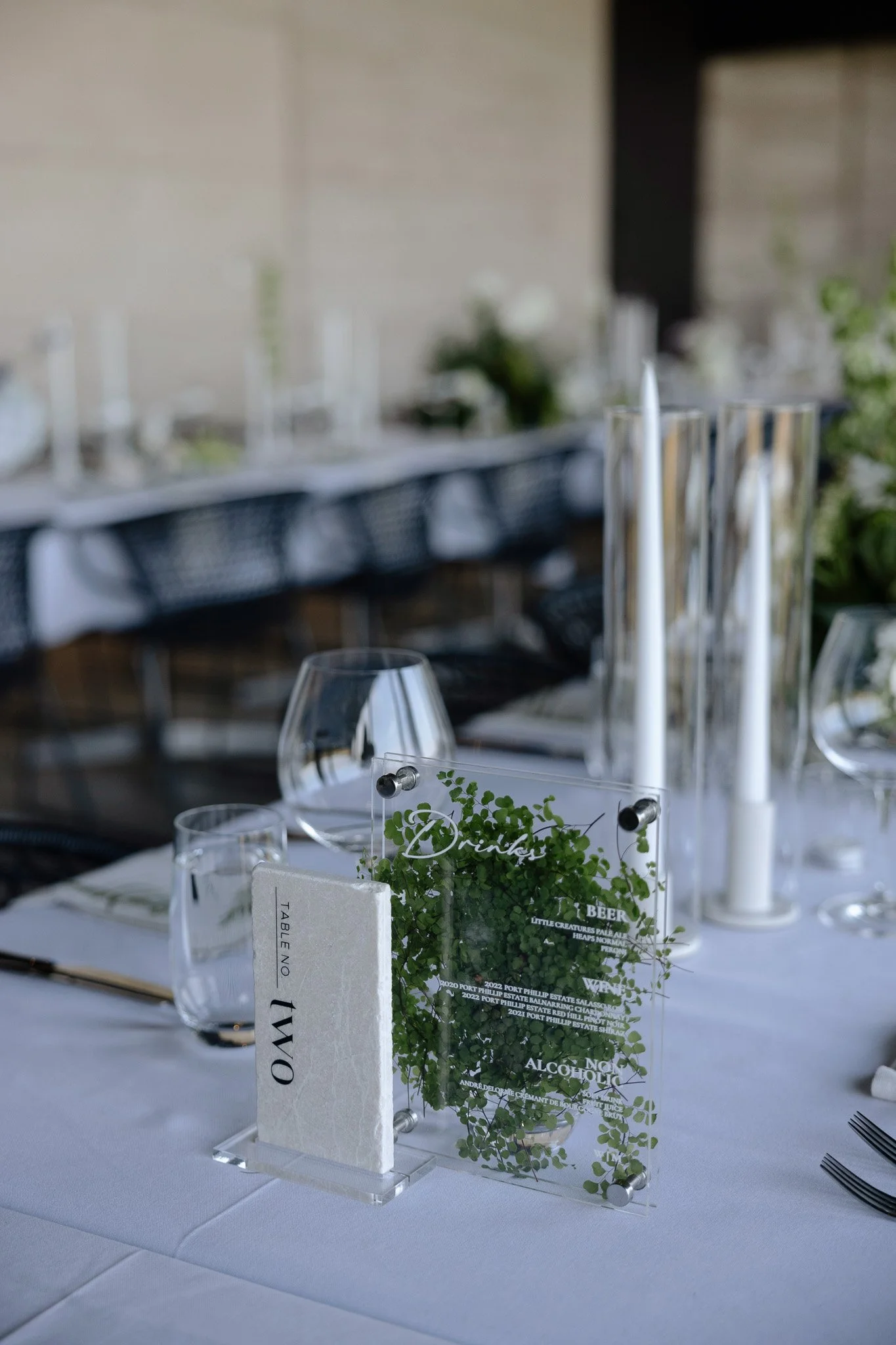 A table centerpiece with a greenery arrangement in a clear glass container, with white candles in tall glass holders around it, and a small decorative sign reading 'Drinks' in a bright, elegant setting.