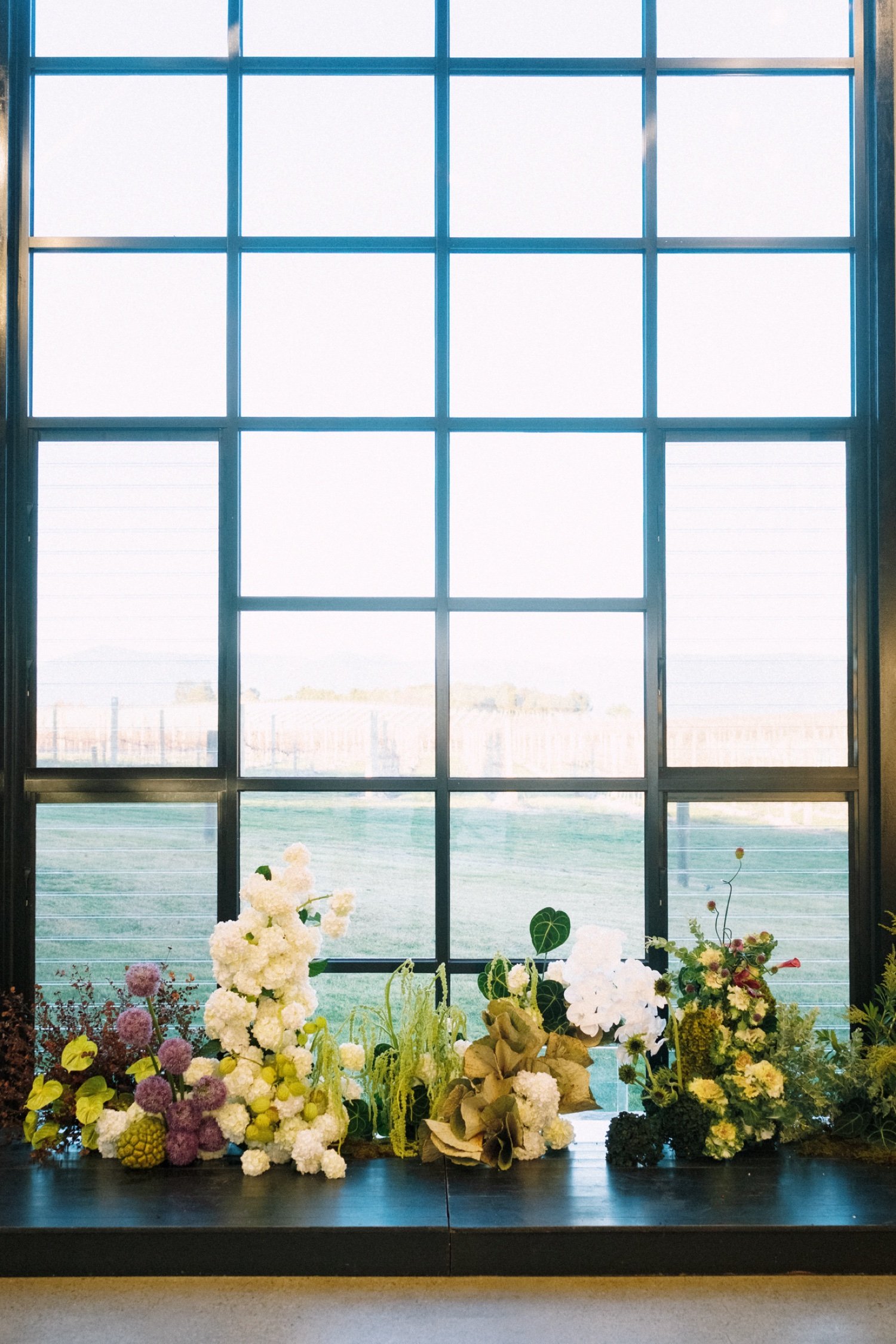 A large window with multiple panes, showing a view of fields or a landscape outside. Below the window, there are various colorful flowers and plants arranged on a dark surface.