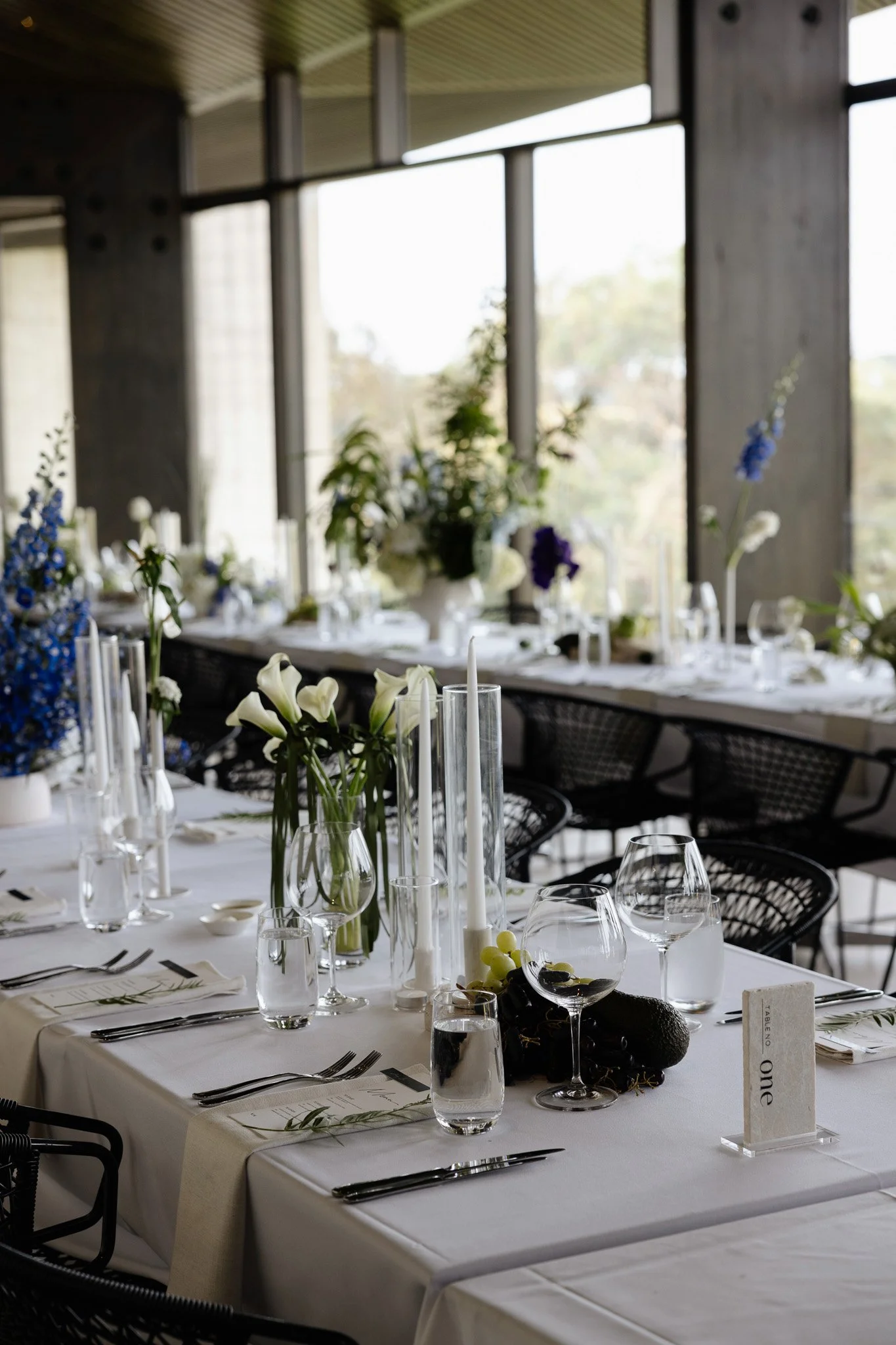 Elegant indoor dining table decorated with white flowers, candles, and glassware, set for a formal event with window views in the background.