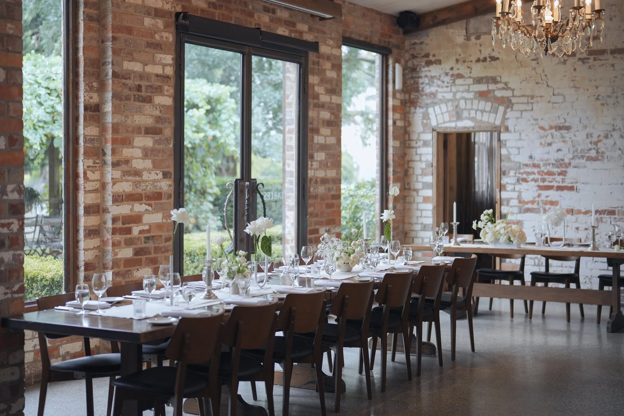 A rustic indoor event space with brick walls, large windows, and a chandelier overhead. The table is set with white tablecloths, floral centerpieces, and glassware, suggesting it is prepared for a formal gathering or celebration.