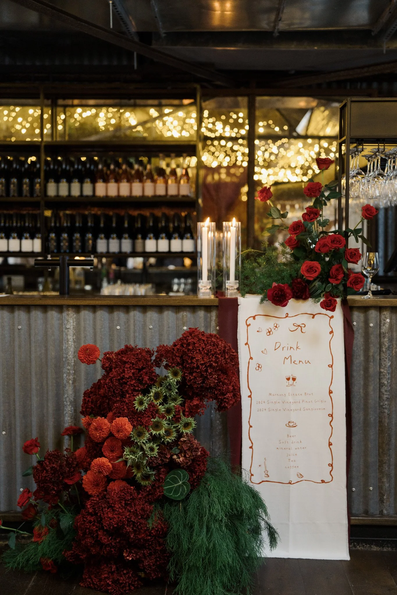 Indoor bar area with a floral arrangement of red and orange flowers and greenery, candles, and a drink menu sign featuring wine and beverages, with shelves of bottles and glasses, and warm string lights in the background.