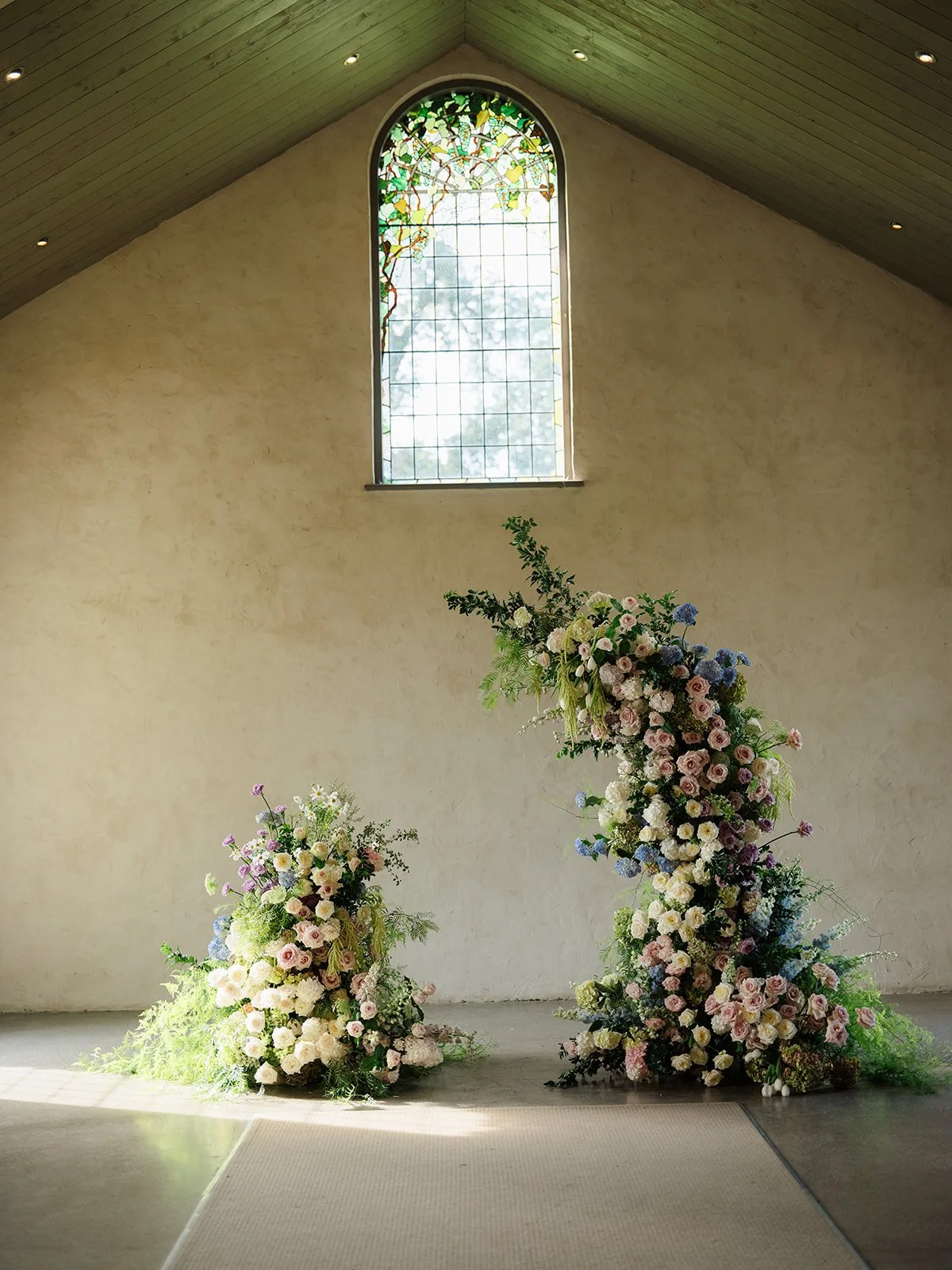 Two tall floral arrangements with pink, white, and purple flowers in front of a beige wall inside a church with a stained glass window and wooden ceiling.