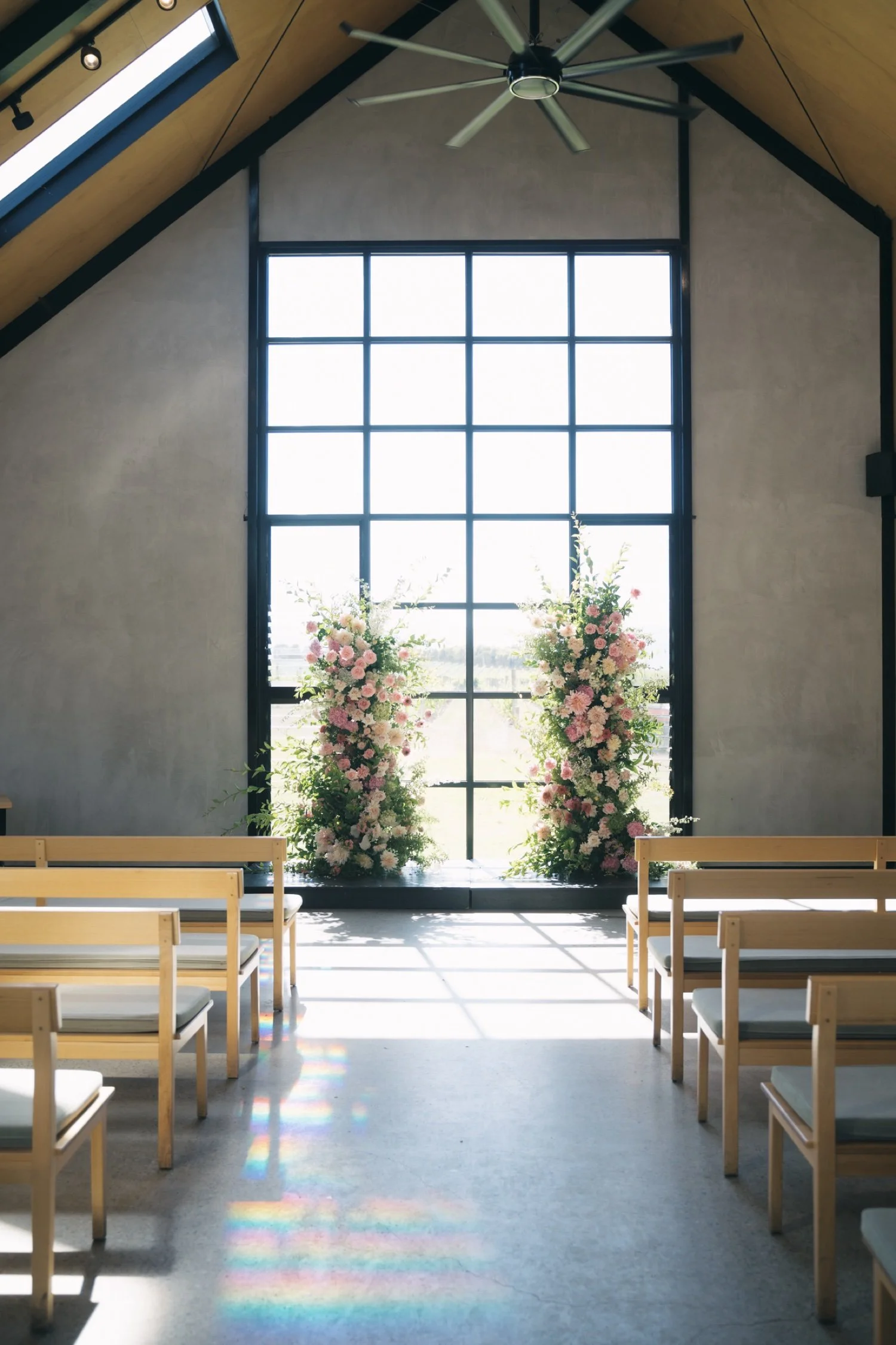 Interior of a church or chapel with wooden benches, a large grid window, and tall floral arrangements of pink and white flowers near the window. A ceiling fan hangs from the wooden ceiling.