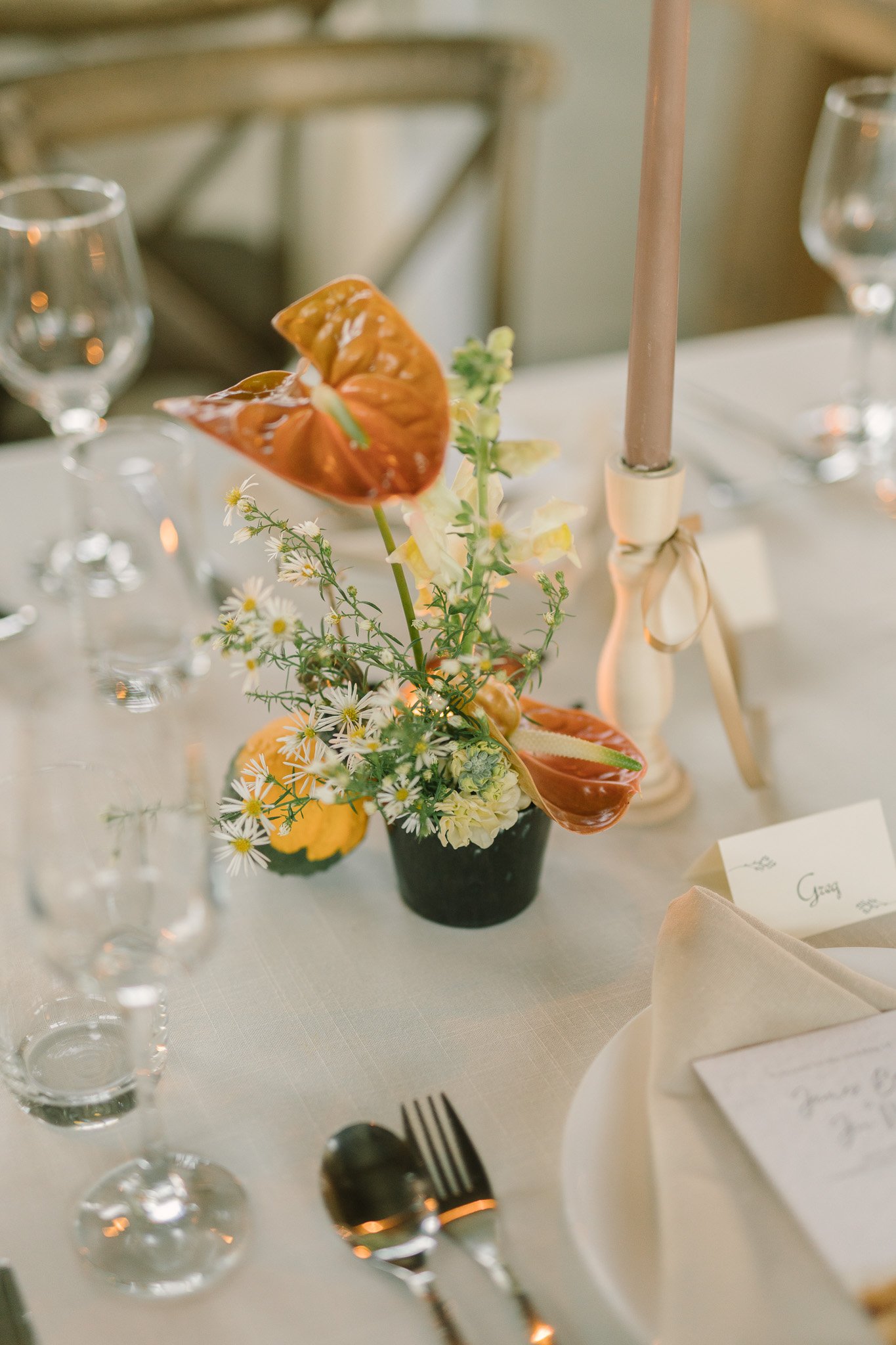 A floral centerpiece with orange anthuriums, white daisy-like flowers, and greenery on a table setting with wine glasses, a candle in a holder, and place cards.