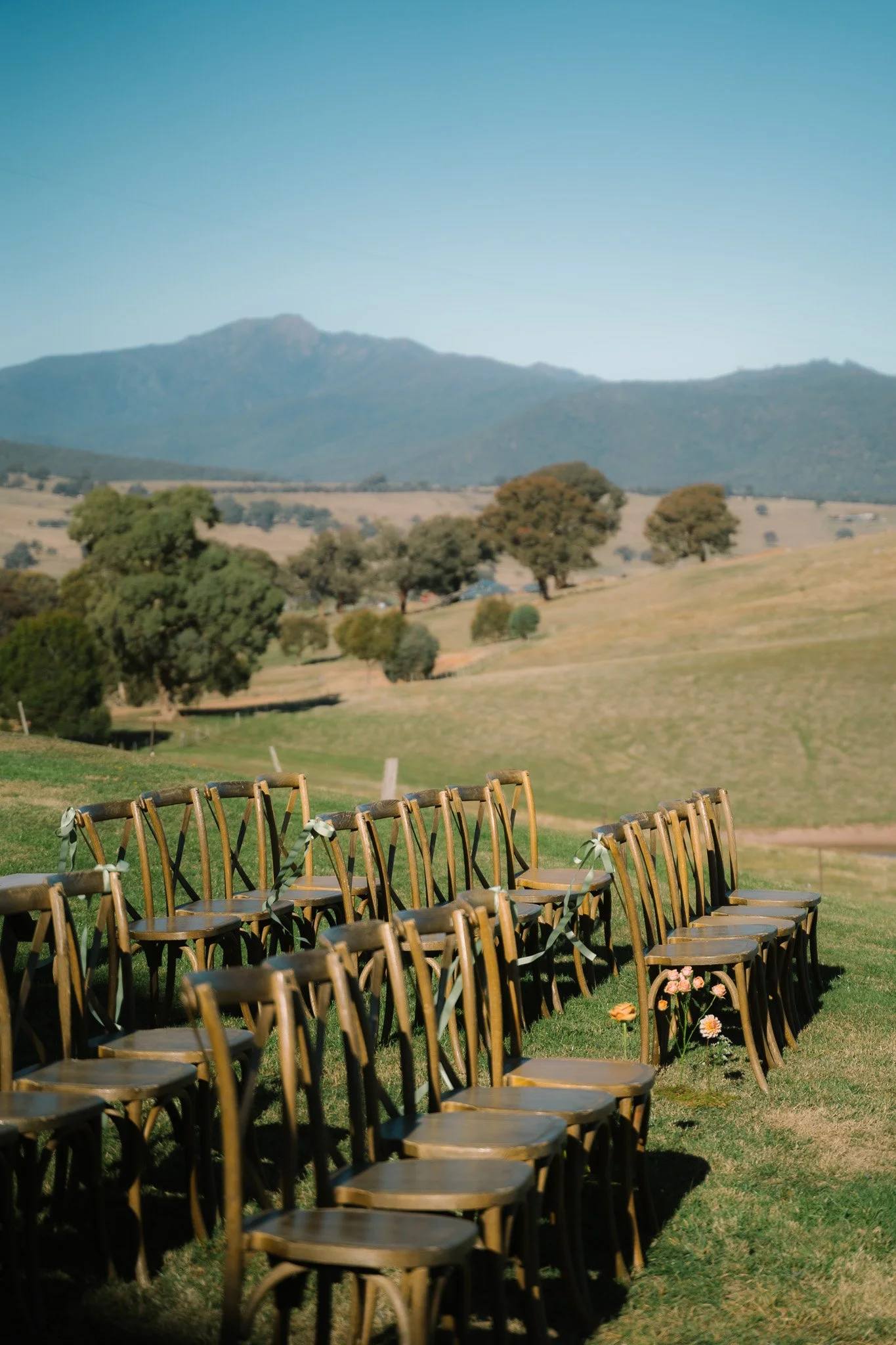 Outdoor wedding setup with rows of wooden chairs on a grassy field, overlooking rolling hills and distant mountains under clear blue sky.