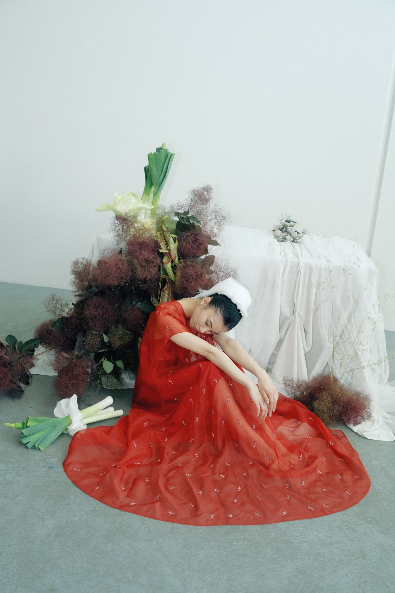 A woman in a flowing red dress sits on the floor with her arms resting on her knees, surrounded by green onions, white flowers, and a floral arrangement. She appears to be introspective or resting in a minimalistic setting with a white backdrop and d