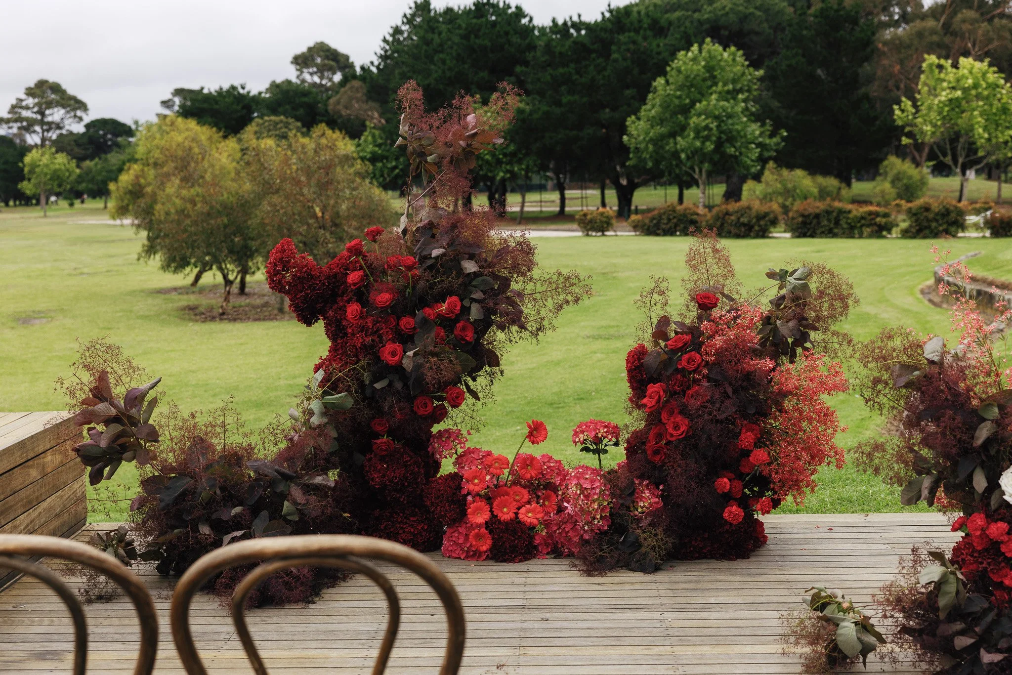View of a park with green grass, various trees, and shrubs in the background, seen from a wooden deck with flower arrangements featuring red and dark purple flowers in the foreground.