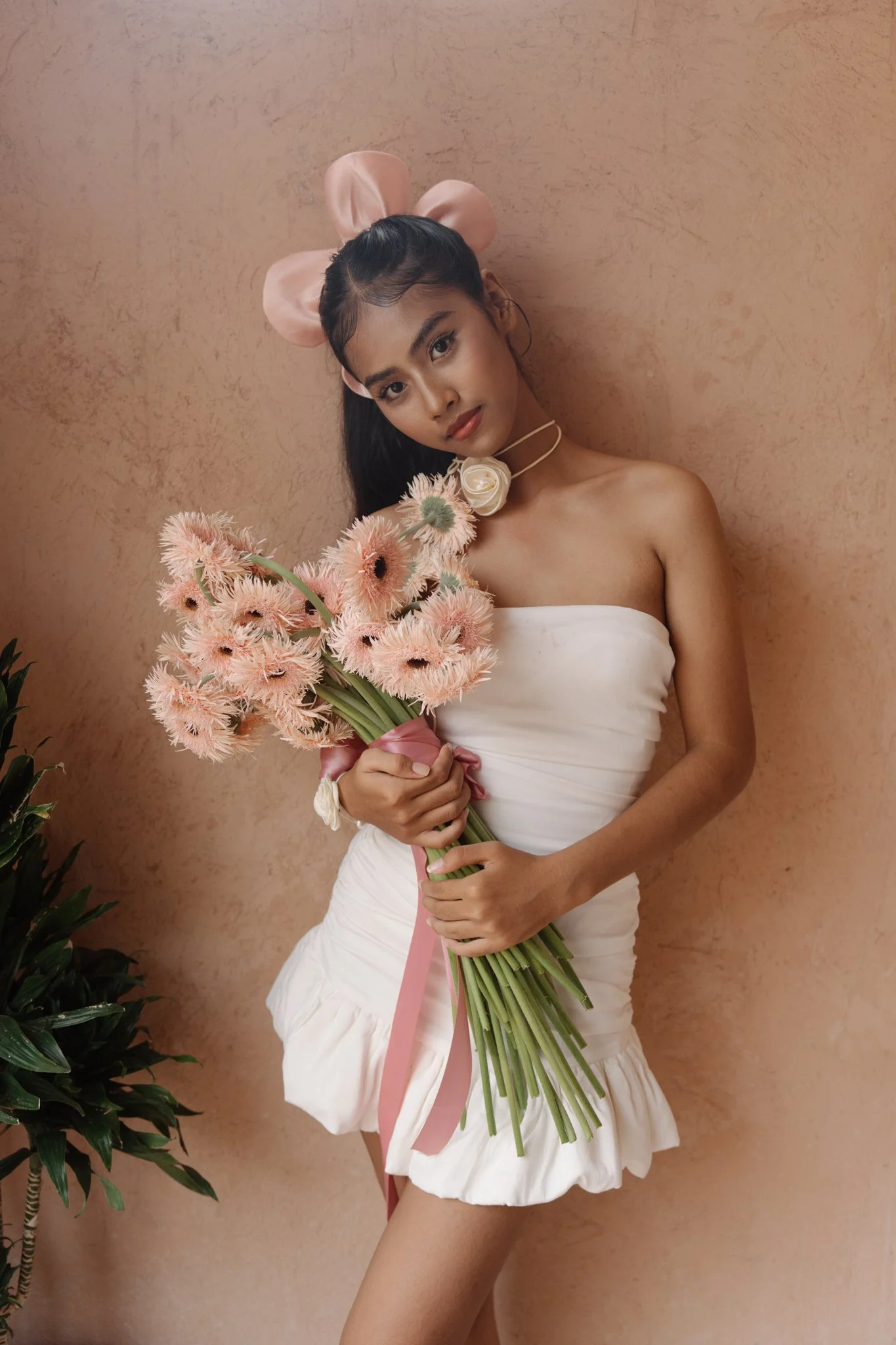 Young woman with long dark hair wearing a strapless white dress, a large pink bow headband, a pink ribbon choker with a large white rose, and holding a bouquet of light pink flowers, standing against a light brown textured wall.