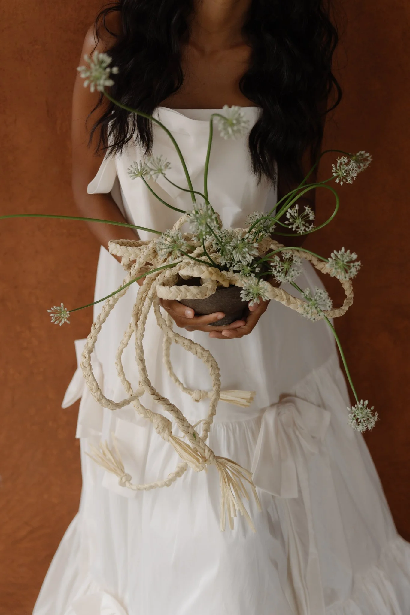 A woman with dark hair wearing a white dress, holding a dark-colored bowl with white flowers and twisted rope decorations, standing against a reddish-brown wall.