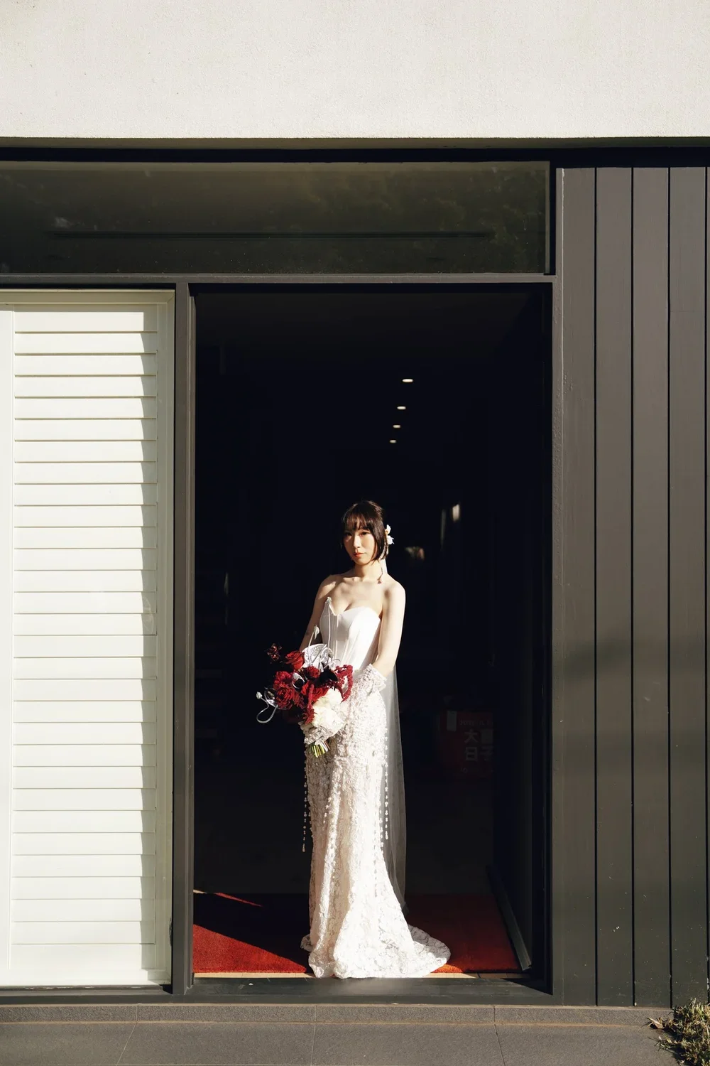A woman in a wedding dress holding a bouquet of red and white flowers standing in a dark doorway with sunlight illuminating her.