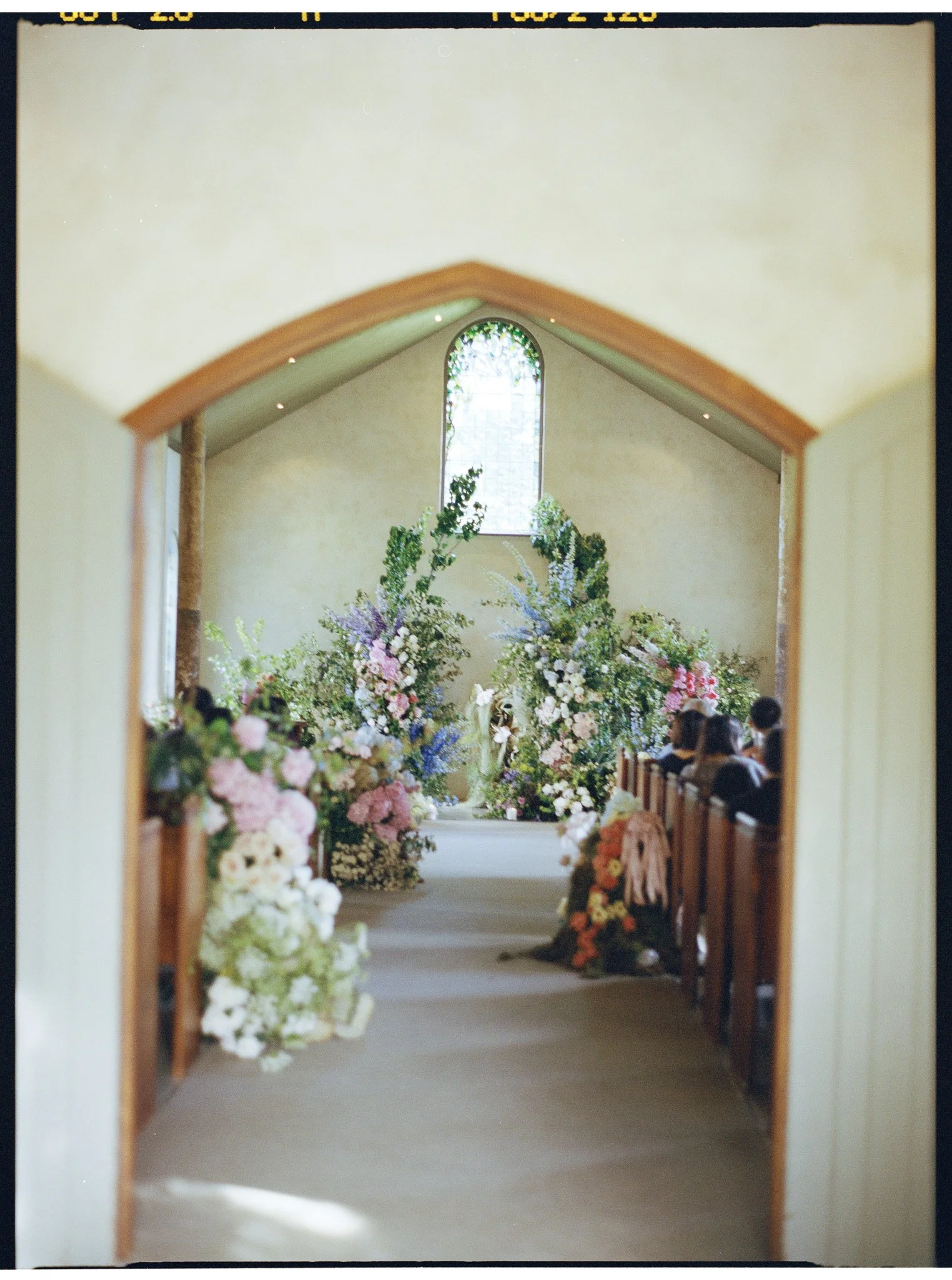 Interior view of a chapel or church aisle decorated with numerous colorful floral arrangements on both sides. There is a large floral display at the end of the aisle near the altar, and a stained-glass window is visible at the far end of the room.