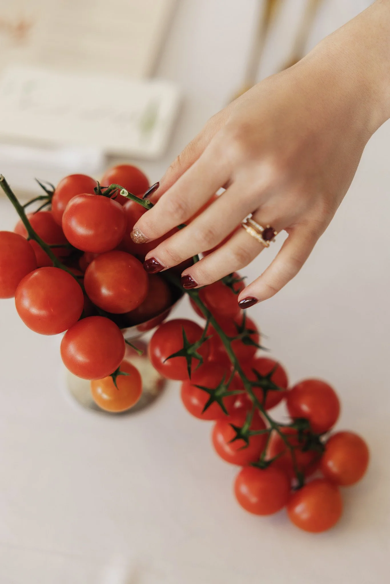 A hand with dark red nail polish and rings touches a bunch of small red cherry tomatoes on a vine, with a blurred background.