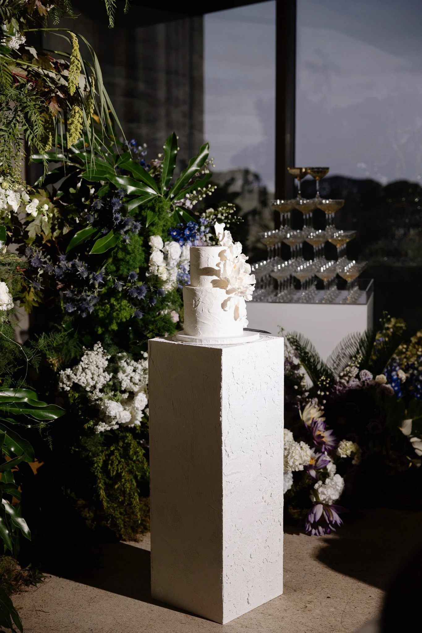 A white, textured, three-tiered cake on a white pedestal in front of a floral arrangement with white, purple, and green flowers and foliage, against a dark indoor background with a window and champagne glasses arranged in a pyramid.
