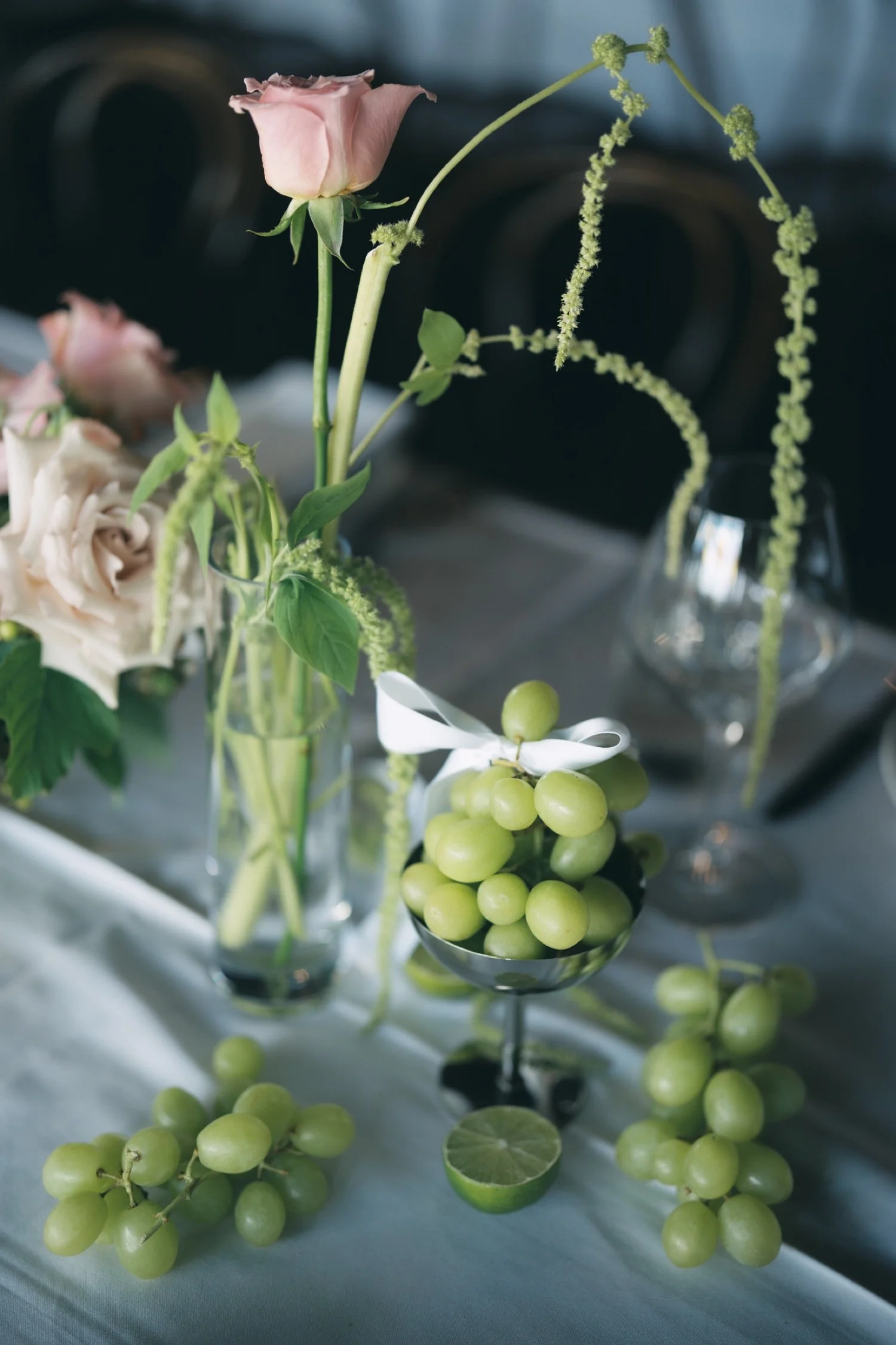 A pink rose, a cream-colored rose, and green grapes in a silver bowl, with some grapes and leaves scattered on a white surface, and a dark background.