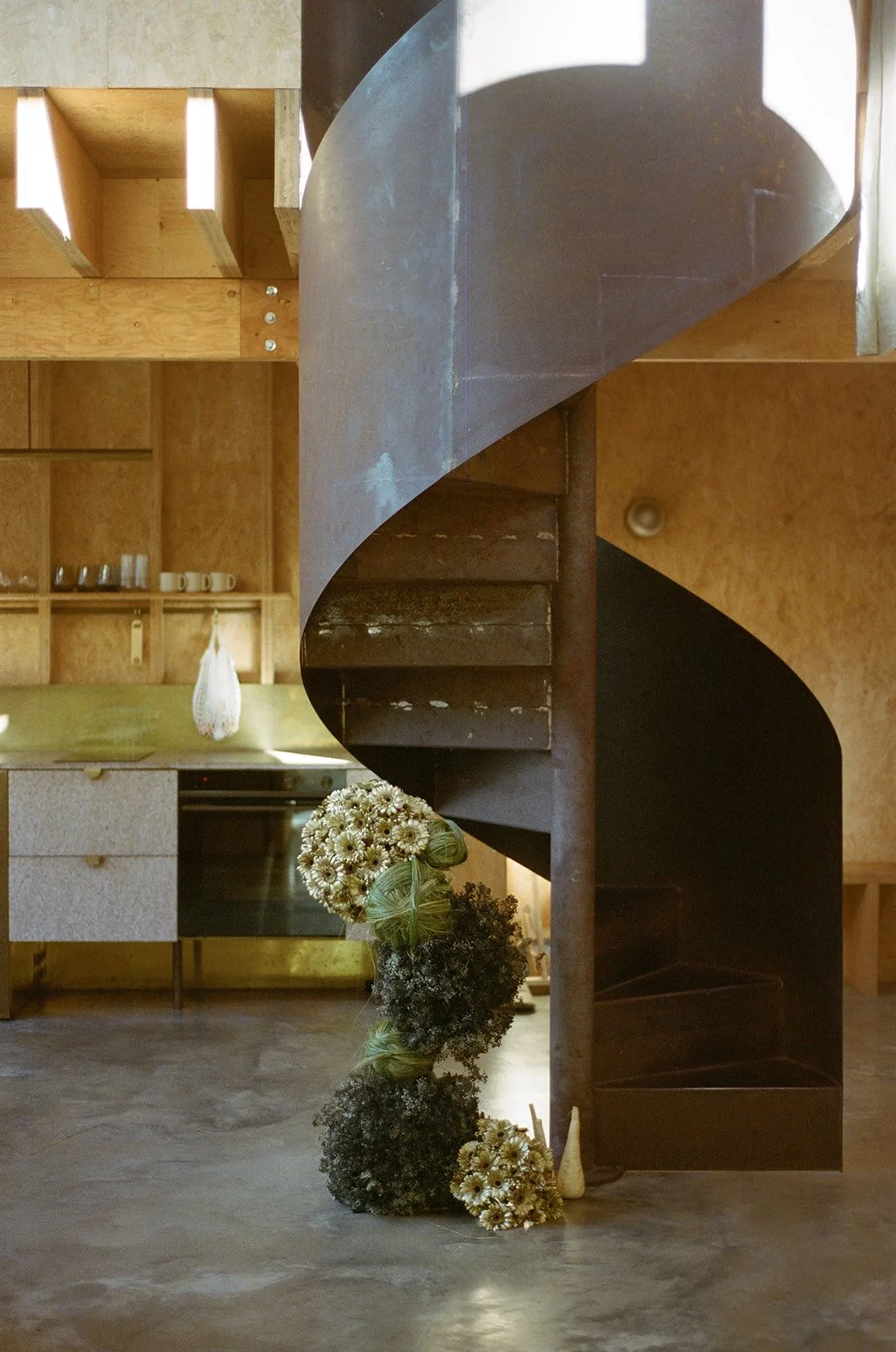 Interior of a rustic kitchen with a wooden cabinet, a stove, and a spiral staircase made of metal leading to an upper level. A large bouquet of white daisies and dark foliage rests at the base of the staircase.