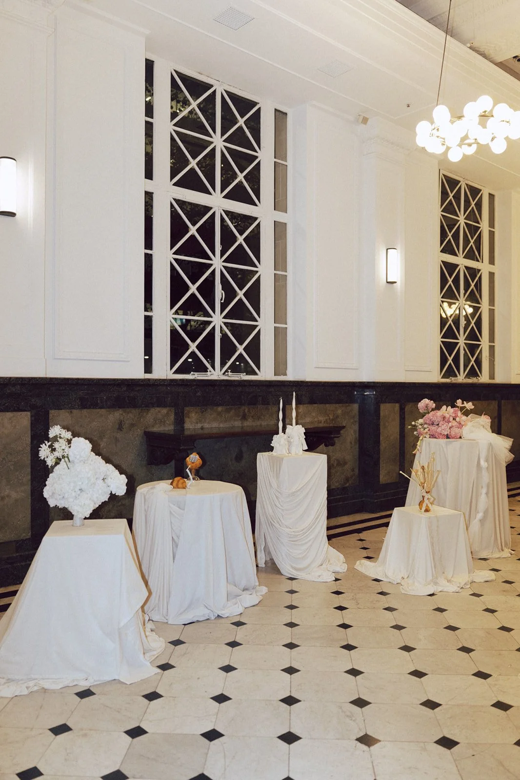 Elegant event decor with white draped tables, floral arrangements, candles, and decorative accents in a bright, luxurious room with large windows.
