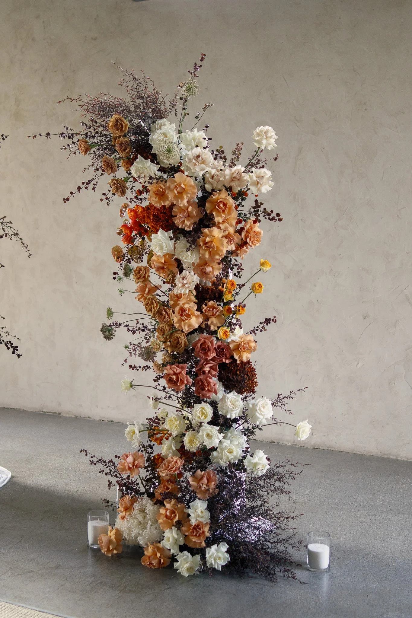 A tall floral arrangement composed of peach, white, cream, and dark purple flowers and foliage, placed on a grey carpeted floor with two white candles in glass holders nearby, against a textured grey wall.
