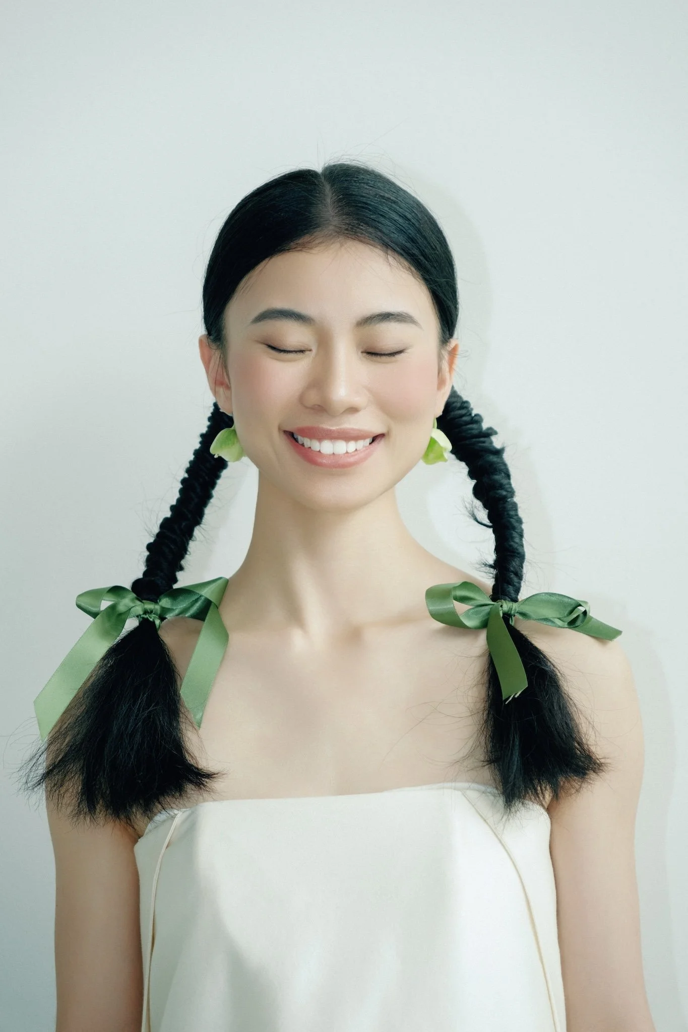 A woman with black hair styled in two braid pigtails tied with green satin ribbons, smiling with eyes closed, wearing a white strapless top, against a plain white background.