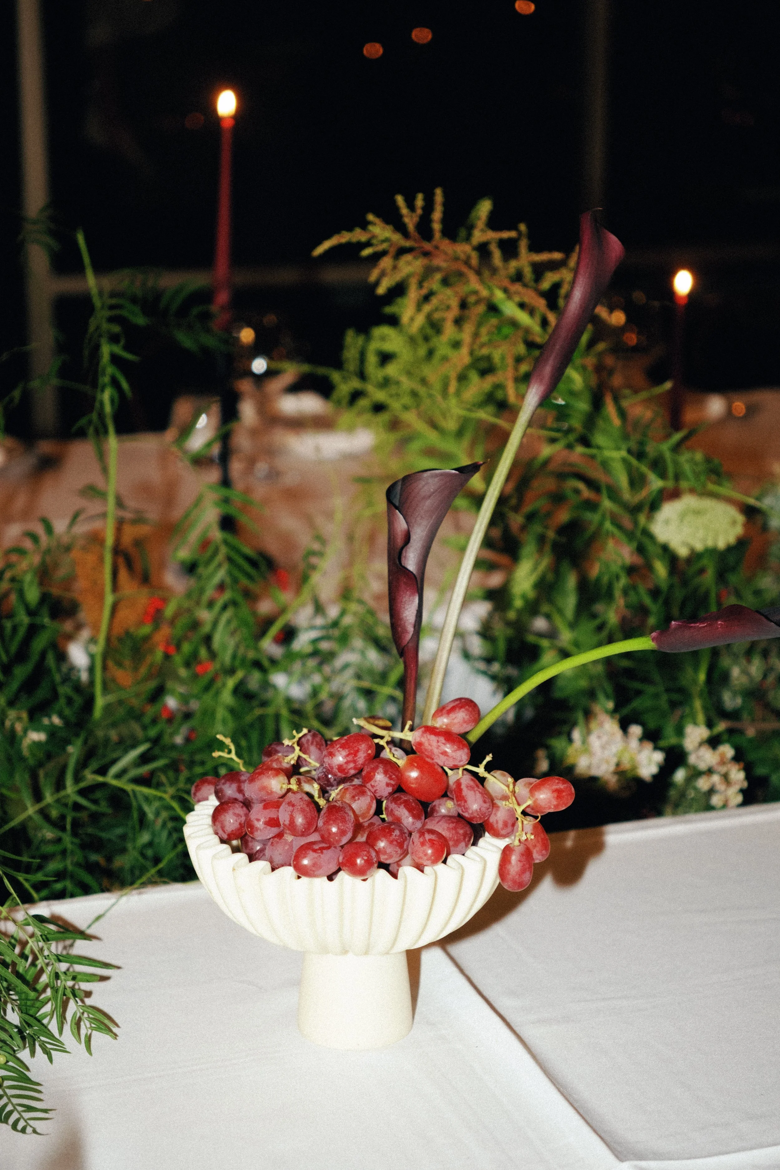 A white pedestal holding a bunch of red grapes in a white bowl, with various green and purple plants and flowers behind it, and some blurred lights in the background at night.