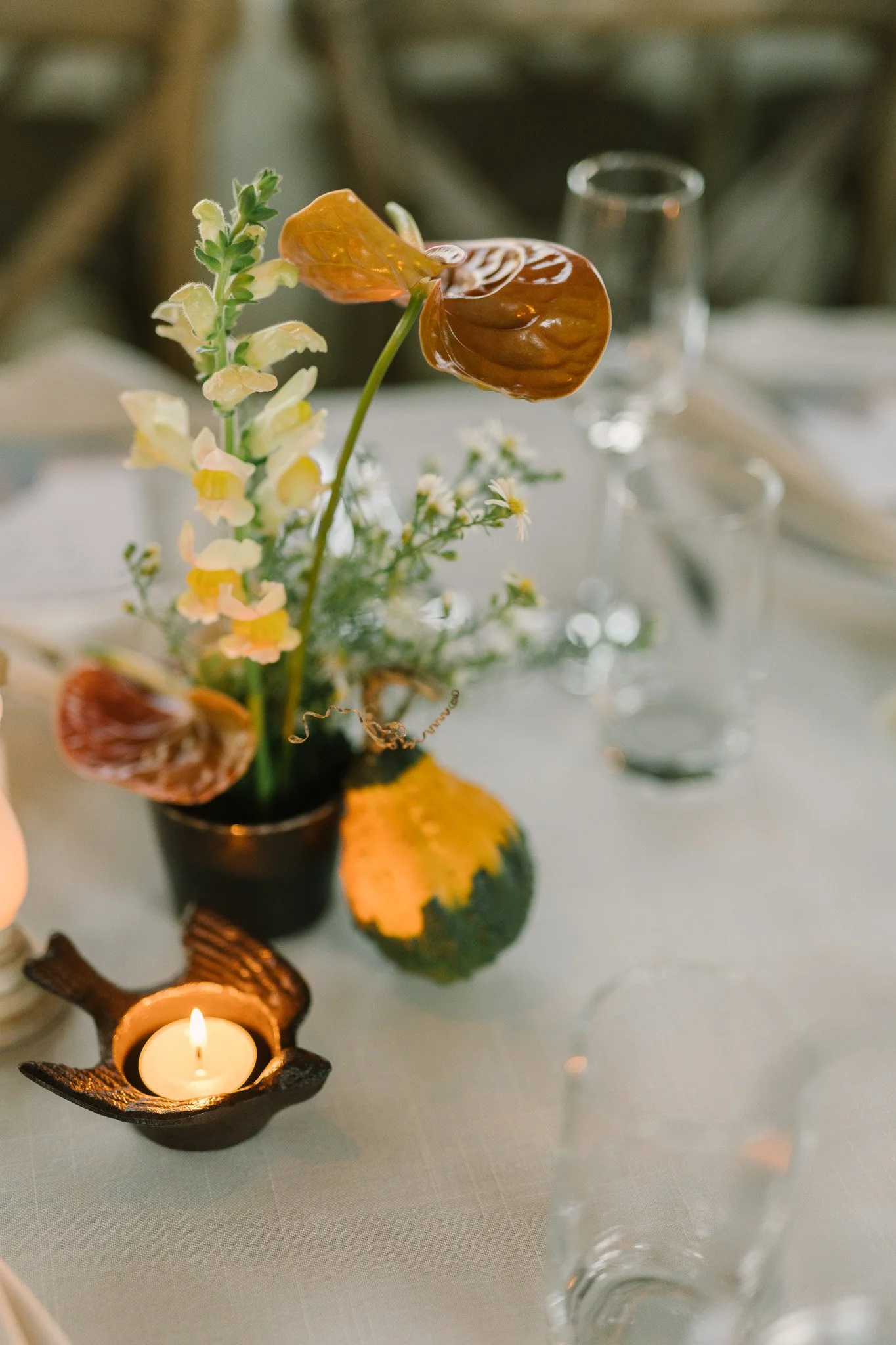 A table centerpiece with flowers and a candle holder with a lit candle, a pumpkin, and a glass of water in the background.