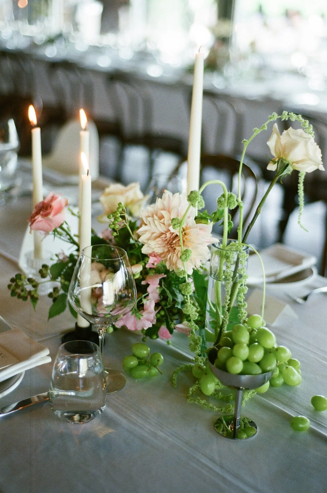 Elegant table centerpiece with white and blush flowers, greenery, and grapes, along with lit candles and glassware.