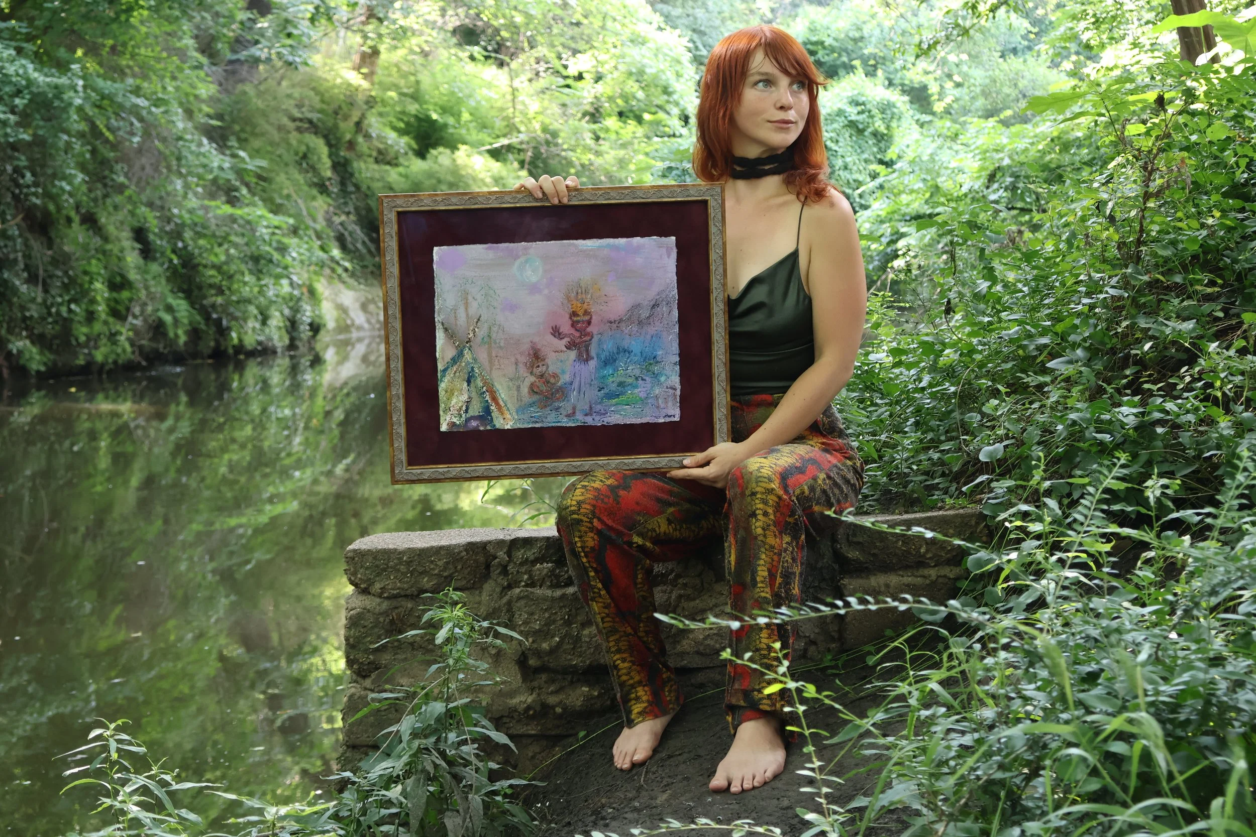 A woman with red hair wearing a black top and colorful pants, sitting barefoot on a stone in a lush green forest near a pond, holding a framed painting of a landscape with a tent, trees, and a moon.